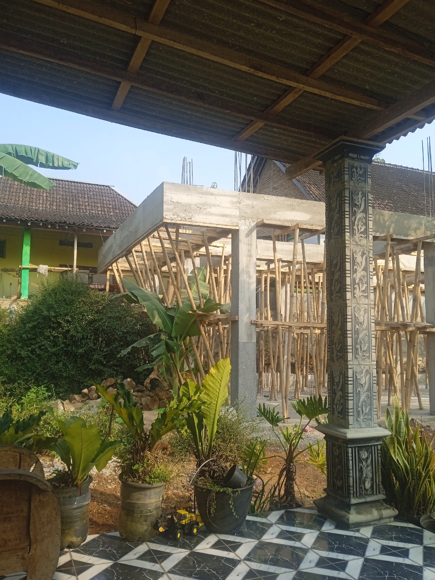This is a ground-level, eye-level, medium shot of an unfinished building structure under construction in Jatisrono, Indonesia. The construction site is supported by numerous bamboo poles, and a concrete structure is visible at the top. The foreground features a tiled patio with a black and white geometric pattern. Several potted plants are placed on the patio, including a large leafy plant in a dark pot, a plant in a recycled plastic bottle planter, and a snake plant. A small yellow toy construction vehicle is positioned on the patio in front of the potted plants. To the left of the patio, a large, weathered wooden barrel is partially visible. In the background, a traditional Indonesian house with a dark tiled roof and a yellow-painted wall is situated behind a dense green bush. The sky is clear and blue, suggesting daylight. A decorative concrete pillar with intricate floral carvings stands prominently on the right side of the patio, framing the construction scene.