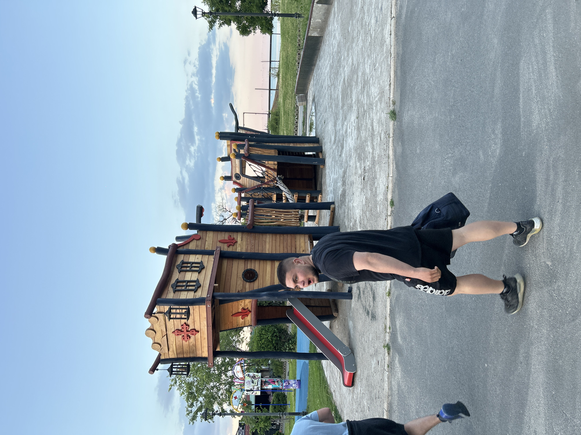 A man wearing a black t-shirt and Adidas shorts is walking on a paved area next to a wooden playground structure with a slide. The playground is in Sozopol, Bulgaria, as indicated by the coastal setting. The sky is light blue with some clouds. Another person wearing a blue shirt is partially visible in the lower left corner of the image.