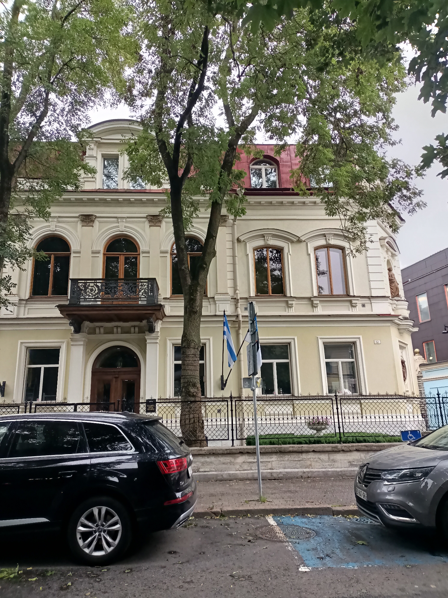 This is a medium-shot, eye-level, outdoor, daylight photograph of a light yellow, three-story building in Tallinn, Estonia, likely located in a historic district. The building features ornate architectural details, including arched windows with dark wood frames, decorative molding, and a wrought-iron balcony. The roof is reddish-brown, and there are trees with green foliage in the foreground, partially obscuring the building. Two flags are visible on flagpoles attached to the building: one appears to be the flag of Finland, and the other is likely the flag of Estonia. In the foreground, two cars are parked on a paved street. The car on the left is a black SUV, and the car on the right is a silver hatchback with a blue-marked parking space. The area appears to be a public street or sidewalk. The overall mood is calm and serene.