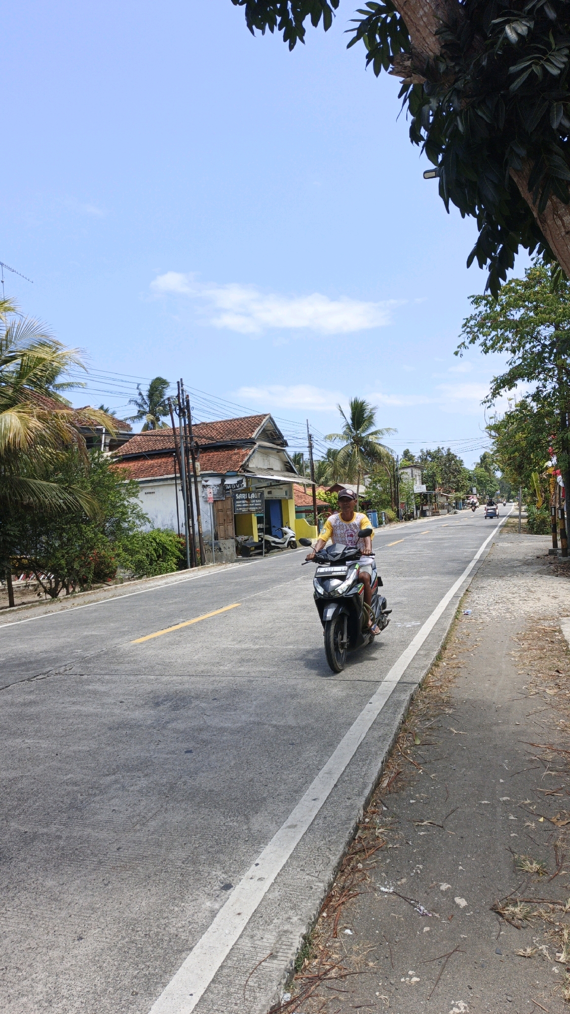 In Cibenda, Indonesia, on a sunny day with a bright blue sky and scattered clouds, a person rides a black scooter down a paved road. The rider is wearing a yellow t-shirt with a graphic print and a baseball cap. The road has a yellow line marking the center and a white line along the right edge. To the left of the road, there are palm trees, a white building with a red tile roof, and another building with a yellow facade. Further down the road, other vehicles, including motorcycles, are visible. To the right of the road is a sidewalk and more trees. The scene depicts a typical day in a tropical environment.