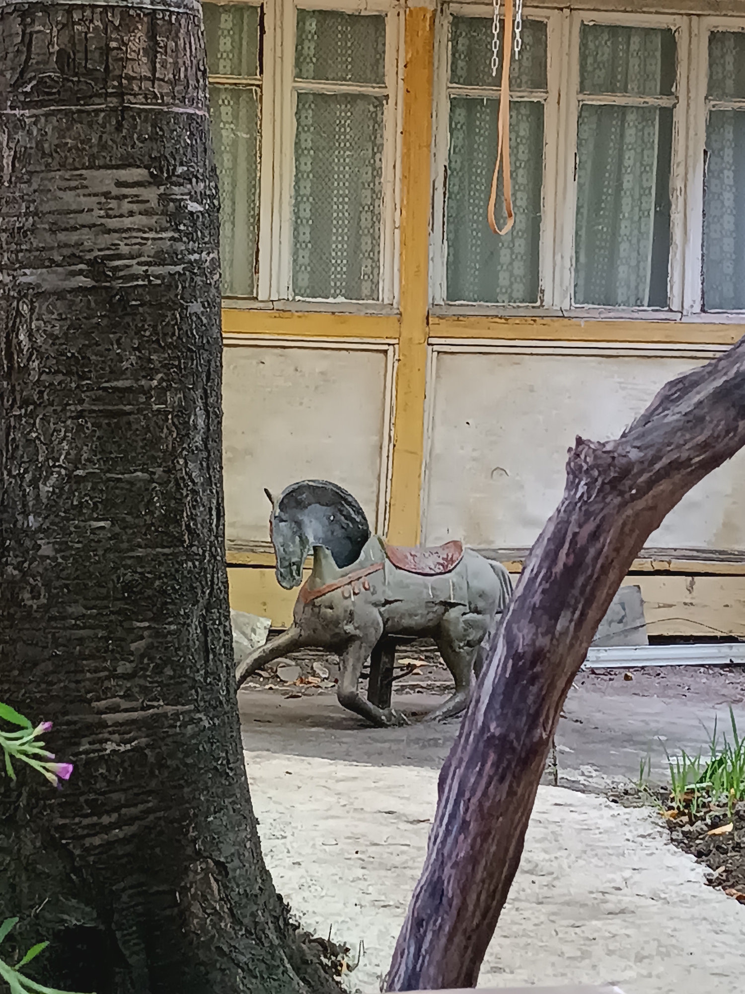 The image depicts a scene with a weathered, stationary rocking horse in an outdoor setting, possibly an abandoned property. The horse, made of an aged material, shows signs of wear and tear, with a detached saddle. It is positioned on a paved surface next to a tree trunk in the foreground. In the background, a building with large windows covered by curtains is visible, suggesting a residential or possibly a former public structure. A chain with a loop hangs from the top of the frame, near the building's windows. The overall atmosphere is one of neglect and decay. This scene is located in Khashuri, Georgia.