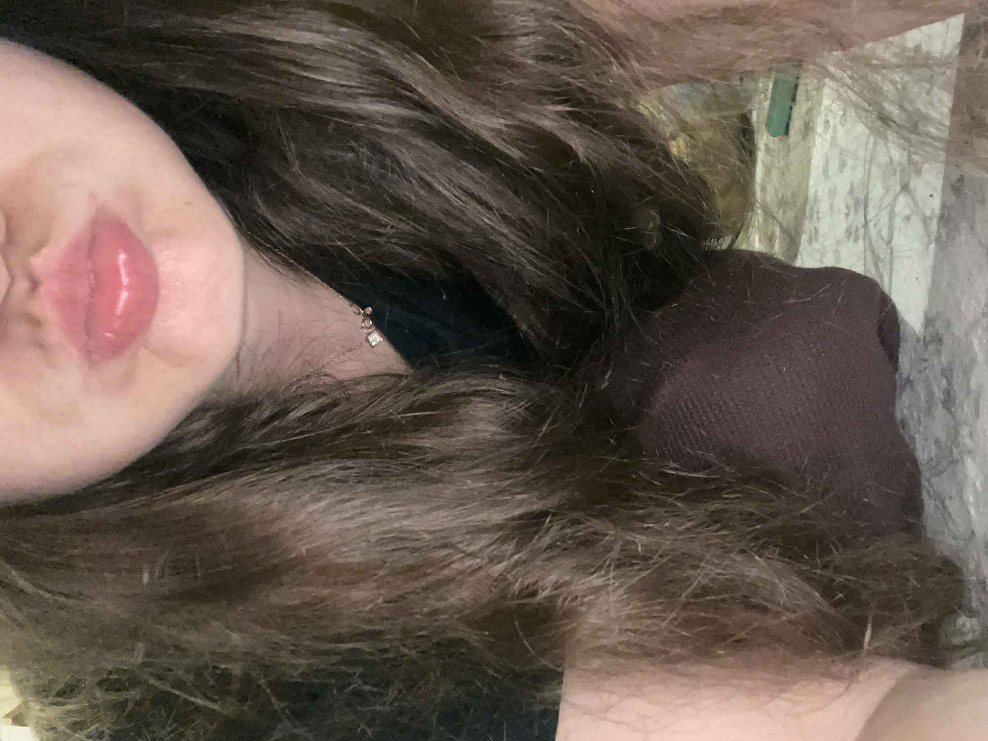 The image, taken in an indoor environment, provides a close-up view of a person's face, focusing on their lips and surrounding area. The individual has long, brown hair that frames their face and falls down their chest. They are wearing a dark-colored shirt and a dark-colored garment, possibly leggings or trousers, visible below their chin. A delicate necklace with a square pendant is also visible, resting on their chest. The background is a textured surface, possibly a floor with a pattern, and a green object is faintly visible to the right. The lighting appears to be soft and diffused.