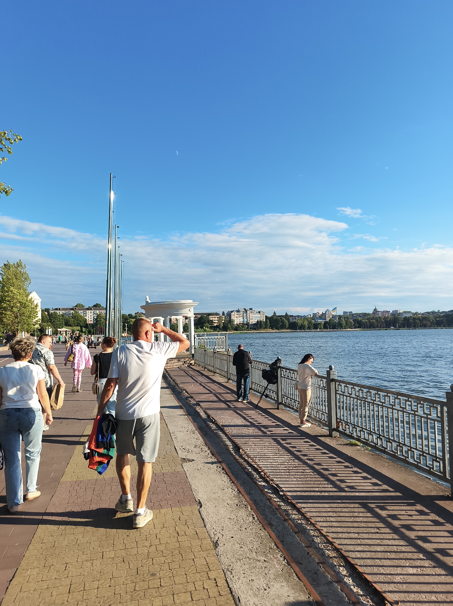 The image depicts a sunny day along the Ternopil Lake promenade in Ternopil, Ukraine. A paved walkway is lined with people strolling and enjoying the view of the water. In the foreground, a man in a white t-shirt and grey shorts walks away from the camera, carrying a bundle of colorful clothing. To his left, a woman in a white t-shirt and blue jeans walks past. Further down the promenade, several other individuals are visible, including a woman in a pink outfit and a couple standing by the railing, looking out at the lake. The lake is calm and reflects the blue sky, dotted with scattered clouds. Across the water, the Ternopil skyline is visible, featuring buildings, trees, and a prominent dome topped with a blue and yellow flag. A decorative white gazebo-like structure is also present along the waterfront. The scene is characterized by its bright lighting and clear weather, suggesting a pleasant day for outdoor activities.