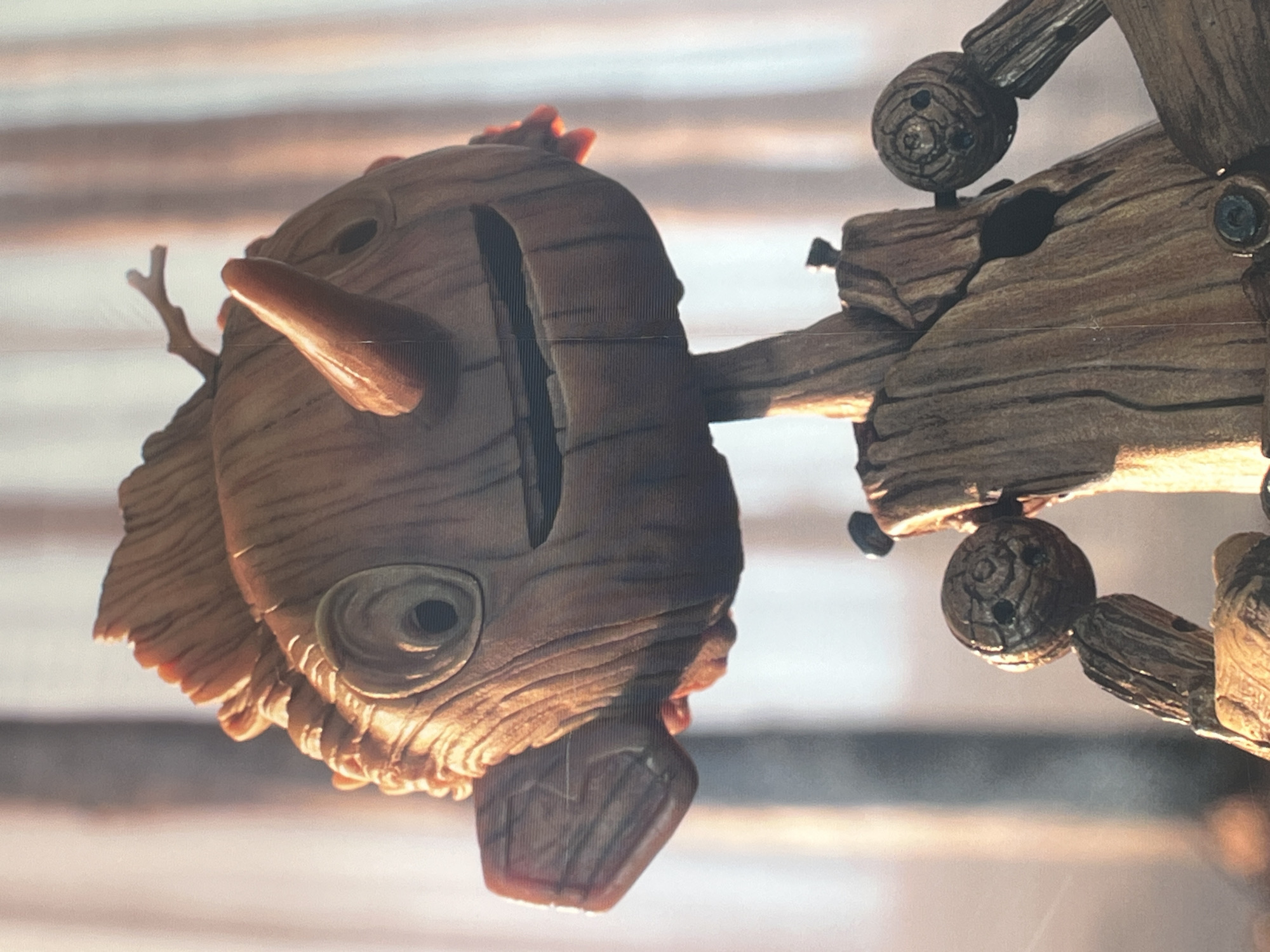 The image shows a close-up of a sculpted head, likely made of wood or a wood-like material, resembling a character. The head features a large, protruding nose-like structure, a deeply set eye, and a slot in the top. The head is connected to a segmented wooden arm or structure, featuring ball joints and various woody segments. The background is blurry and indistinct.