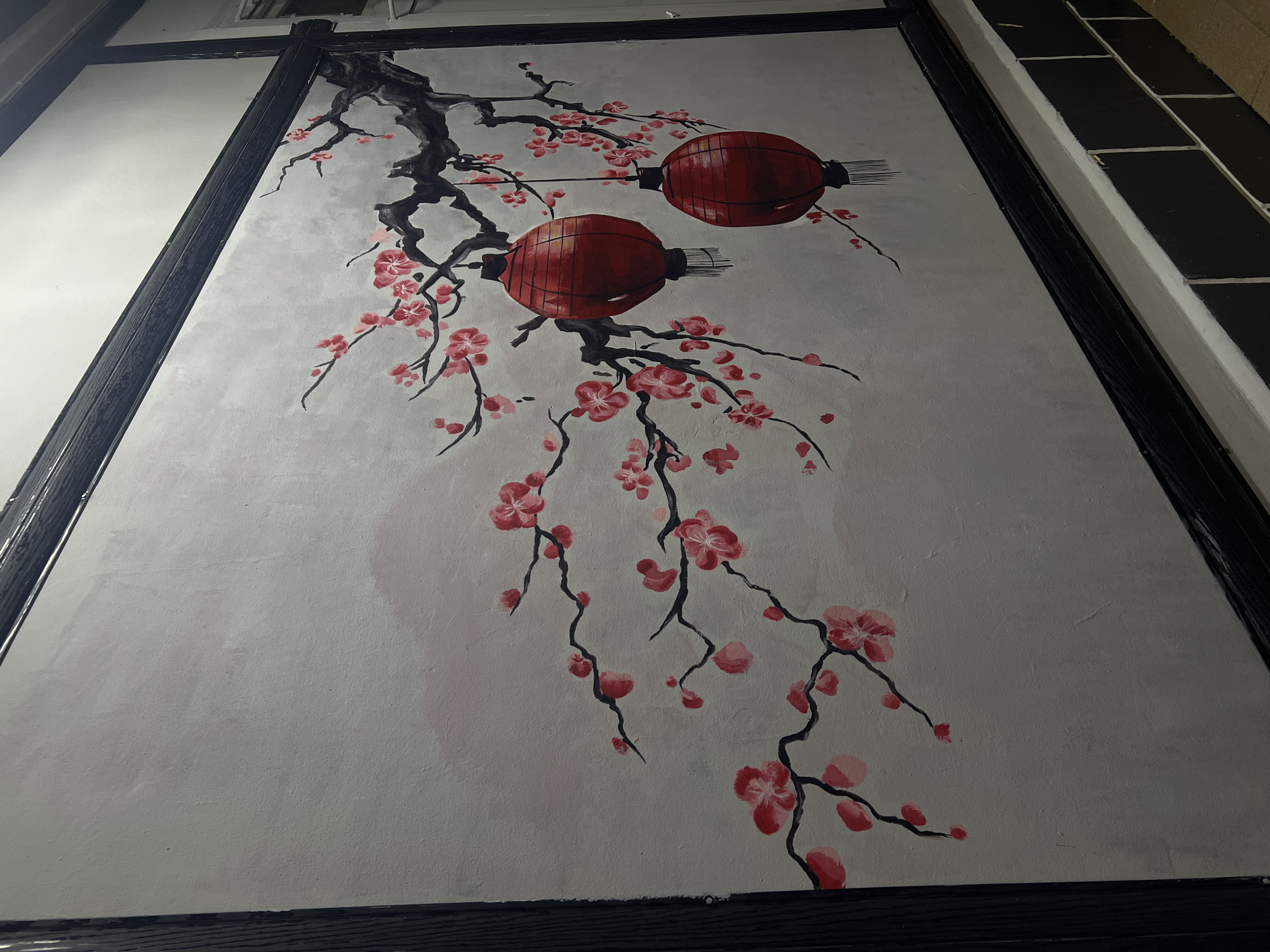The image shows a painting on a wall depicting a cherry blossom branch with two red lanterns hanging from it. The painting is framed by a dark border and is mounted on a light-colored wall. The lower portion of the image shows a tiled wall and a ledge. The scene is located in the city of Shymkent, Kazakhstan, potentially in a restaurant or an establishment with Asian-themed decor. There are no people or animals visible in the image. The style of the painting is traditional, with thin black branches and vibrant pink blossoms. The lanterns are round and red with black accents, and they appear to be illuminated, casting a soft glow. The overall impression is one of serenity and cultural richness.