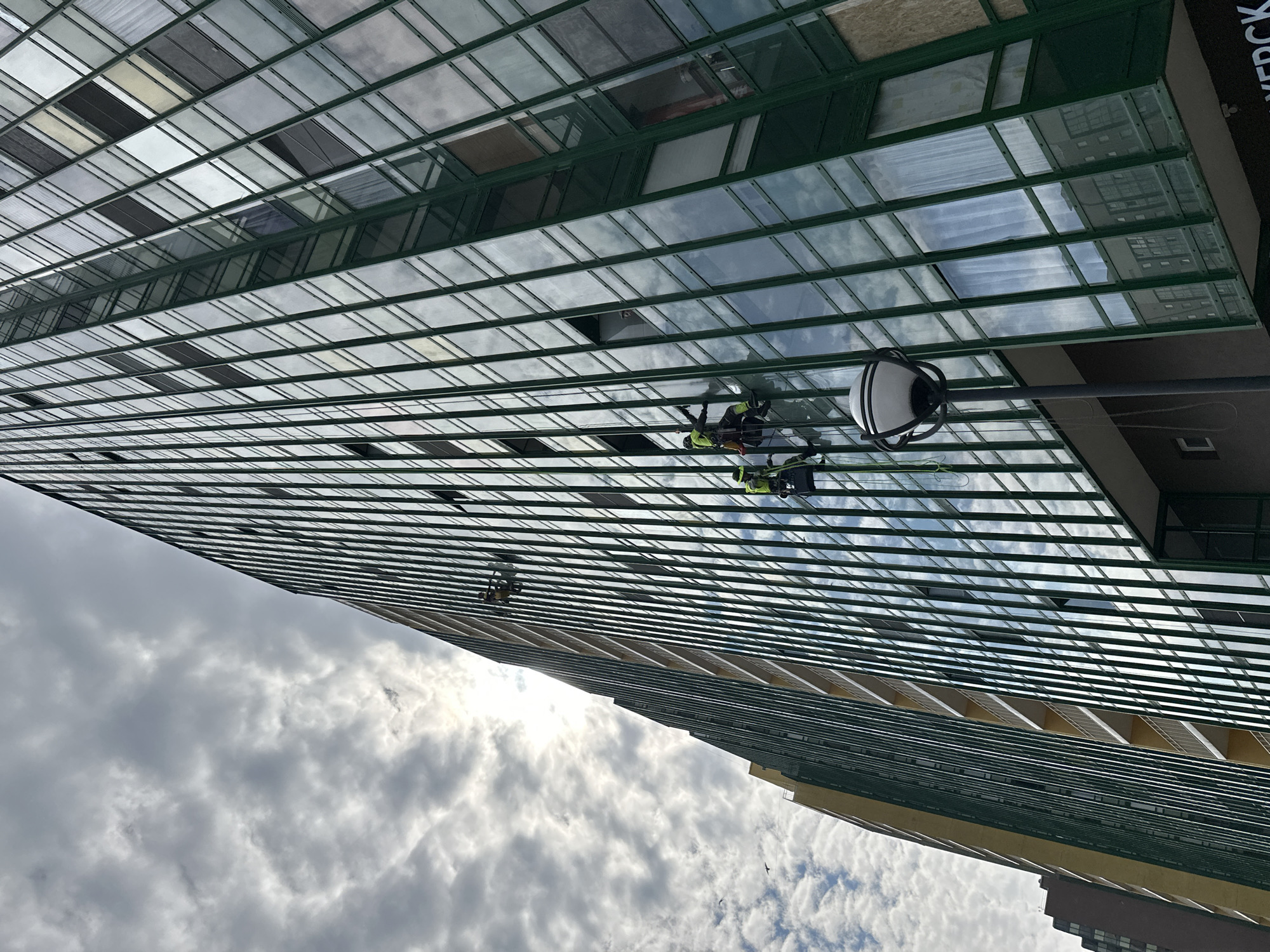 The image depicts a tall, modern building with a grid-like facade of glass windows. Two window washers are visible, suspended by ropes, cleaning the windows. The building's structure features green horizontal and vertical lines. The sky is cloudy. The building is presumably in Murino, Russia, based on the context provided. The building also has a lamp.