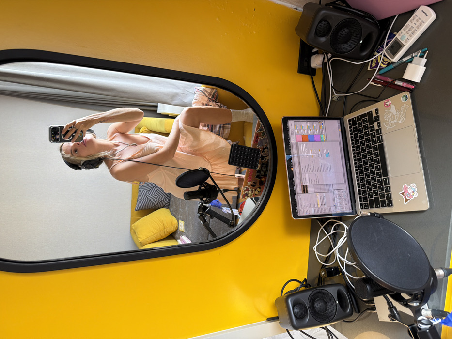 The scene depicts an indoor environment, viewed from an elevated perspective above a desk. A blonde female individual is reflected in a large, black-framed mirror that dominates the upper half of the image. She wears over-ear headphones, a light peach-colored spaghetti strap dress, and white socks. She is holding a dark-colored smartphone, pointing it towards herself, indicating a selfie or video recording. A silver ring is visible on her left ring finger, and a small tattoo is present on her left forearm. She appears to be sitting or kneeling, with one knee bent. Behind her, within the mirror's reflection, is a yellow sofa with several yellow pillows and one black-and-white patterned pillow. A microphone on a stand with a pop filter is positioned near her, alongside a small black MIDI controller or grid device. The reflected background consists of a white wall.
In the foreground, a dark grey desk holds a workstation setup. A silver Apple MacBook laptop, adorned with stickers including a pink character and a winged cupid, is open. Its screen displays a digital audio workstation (DAW) interface, likely Ableton Live, showing multiple tracks and controls. Two black studio monitor speakers flank the laptop. A black condenser microphone with a pop filter is mounted on a stand on the left side of the desk, surrounded by various tangled white and black cables. To the right of the laptop, a black power strip, two pens (pink and green), a white USB-C charging adapter, and a white Daikin air conditioner remote control displaying "27" are arranged. The wall immediately behind the desk is painted a vibrant yellow. The MacBook's keyboard prominently features a Thai layout, definitively placing the location in Bangkok, Thailand.