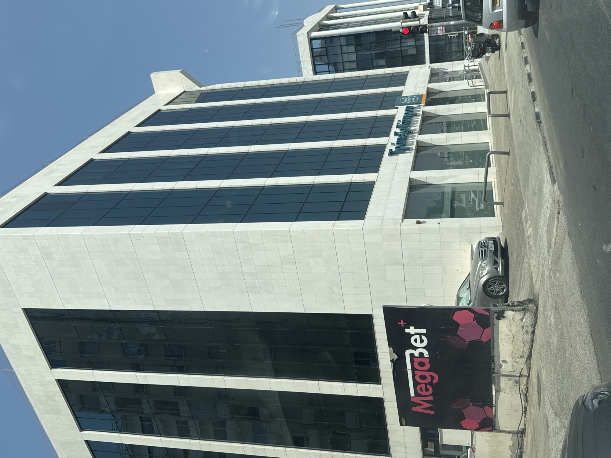 In Limassol, Cyprus, this street-level, outdoor photograph captures a modern, multi-story building with a facade of white stone and dark glass windows. On the left side of the building, a large, black billboard with the bright pink and white "MegaBet" logo and hexagonal pink graphics is prominently displayed. To the right of the MegaBet billboard, a silver sedan is parked. The building's ground floor features large glass windows, one of which displays the text "Τράπεζα Κύπρου" (Bank of Cyprus) in green lettering. Adjacent to the building, a traffic light with a red signal is visible, along with a motorcycle parked on the sidewalk. The scene is set against a clear blue sky.