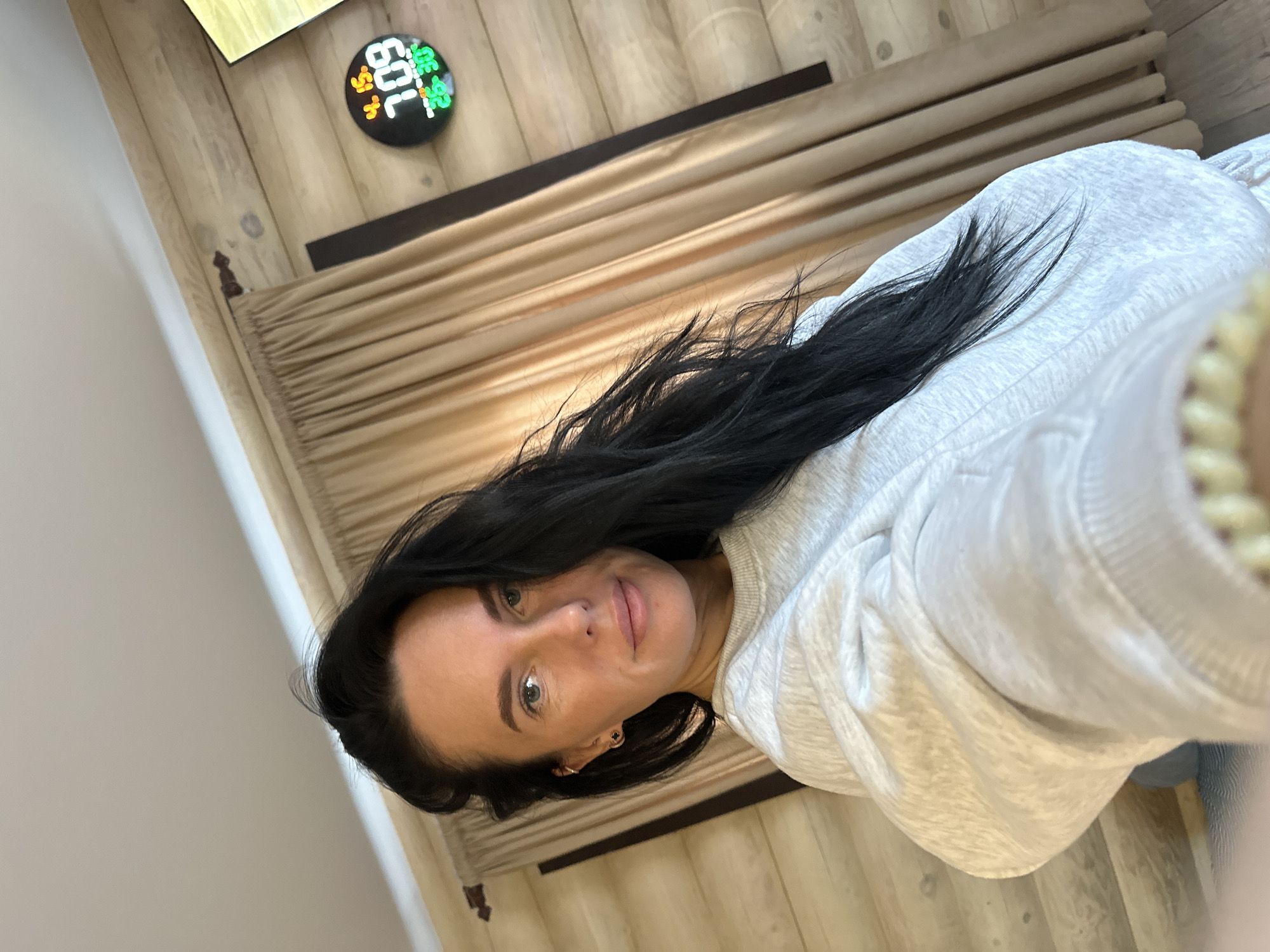 A woman with long, black hair and fair skin is pictured from a slightly high angle. She is wearing a light gray sweatshirt. In the background, there is a wood-paneled wall and a beige, horizontally-striped headboard or wall covering. A circular device with illuminated numbers, possibly a thermometer or clock, is mounted on the wall. The location is inferred to be Ольховка, Russia. There are no visible actions or interactions.