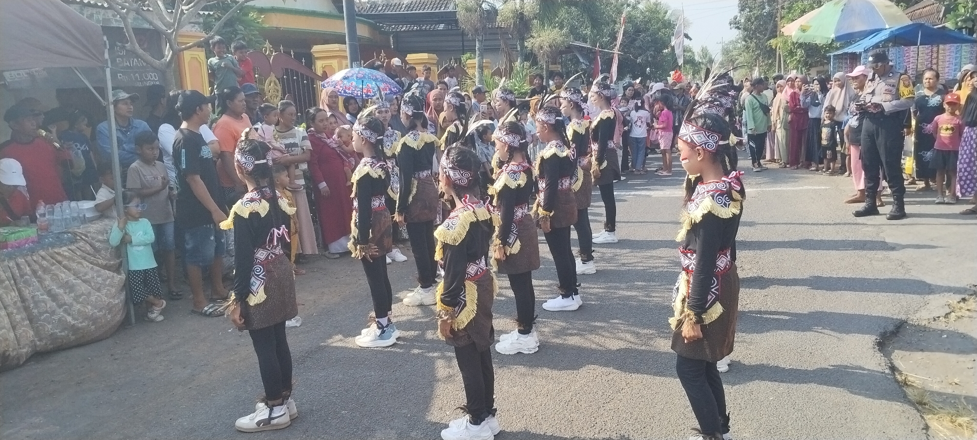 This image captures a cultural event taking place on a street in Yosowilangun Kidul, Indonesia. In the foreground, a group of young girls dressed in traditional attire with feathered headbands and patterned skirts stand in formation, appearing to be part of a parade or performance. They wear black tops with decorative elements and brown skirts. Behind them, a crowd of onlookers, including adults and children, observe the proceedings. Various stalls or temporary structures, possibly for vendors, are visible in the background, along with buildings and trees. A police officer in uniform is also present, overseeing the event. The atmosphere appears festive and community-oriented, with people gathered to watch the cultural display. The overall scene depicts a vibrant street event in a local Indonesian community.