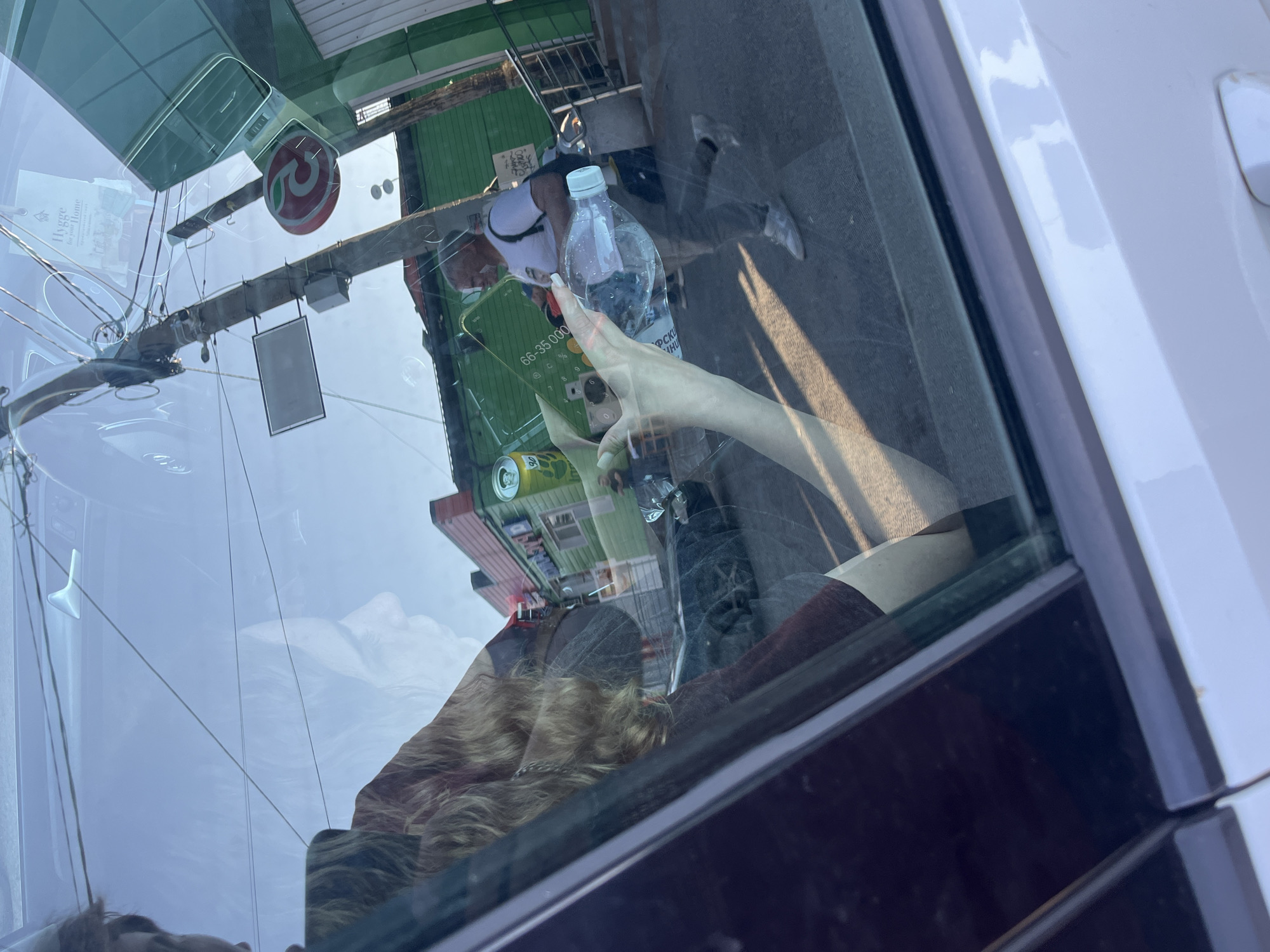 This image, captured in Vasilyevo, Russia, presents a complex scene viewed through a car window, with reflections overlaying the exterior environment. The foreground is dominated by the interior of a white car, with the edge of the driver's side door visible. A person with blonde hair, seen in profile, is reflected in the glass, their face partially obscured.
Outside the car, a street scene unfolds. A utility pole with numerous wires stands prominently, adorned with various signs and electrical equipment. One sign features a red circular logo with a green apple motif. A green building with a pink roof is visible in the background, possibly a shop or market.
Several individuals are present in the reflected outdoor scene. A man in a white t-shirt and grey pants is walking, carrying a backpack and a clear plastic bottle of water. Another person, partially visible in the lower right, appears to be standing. The hand of a person inside the car is holding a smartphone with a green case, and an open can of soda with yellow branding is also in view. The ground appears to be paved. The lighting suggests it is daytime, with bright sunlight casting shadows.