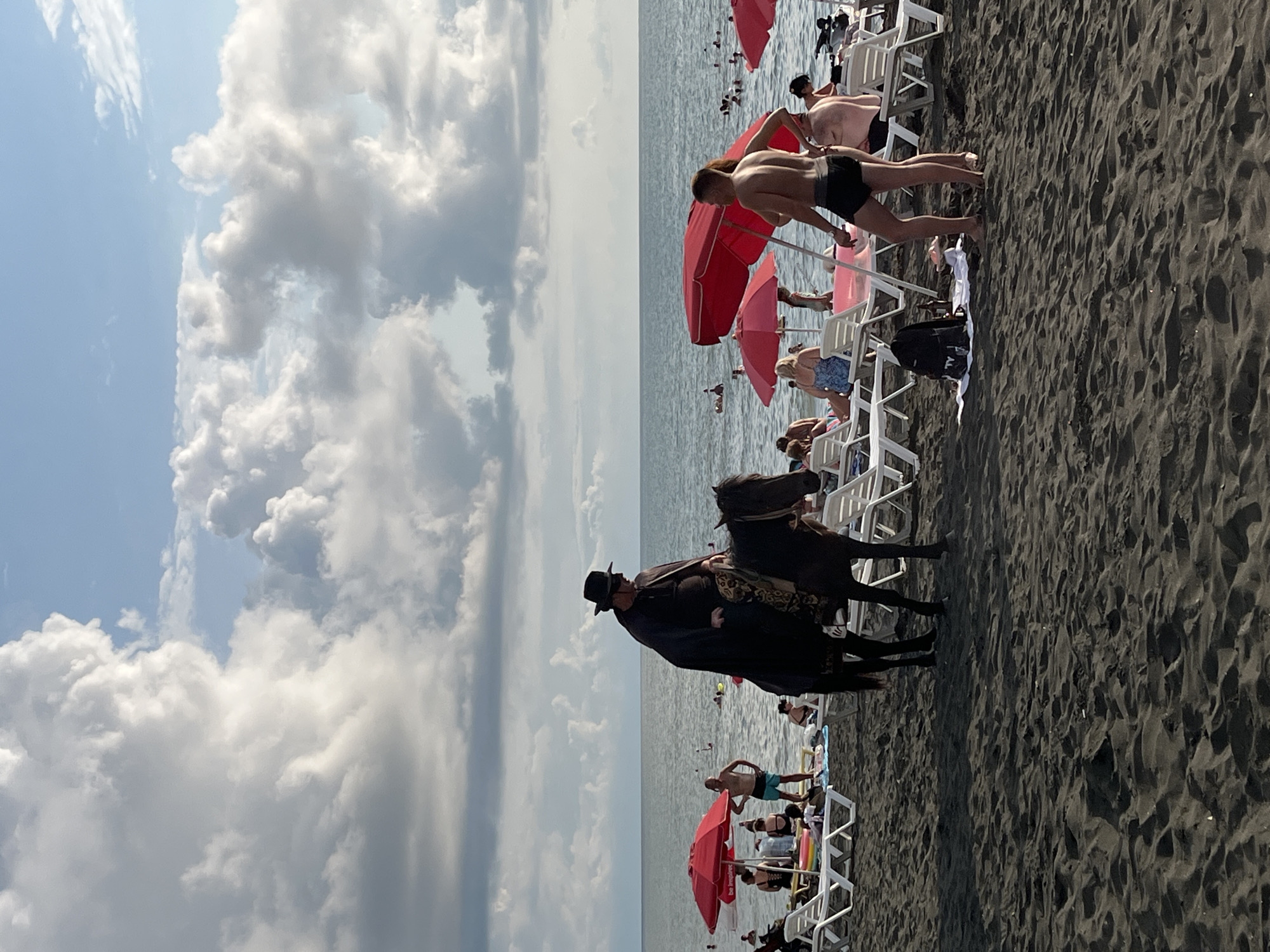 A man dressed in a black cloak and hat rides a dark horse on a black sand beach in Ureki, Georgia. The sea is calm with a few people swimming in the distance. Beach chairs and red umbrellas are set up on the sand. Several people are visible, some relaxing on chairs, others walking or in the water. The sky is partly cloudy.
