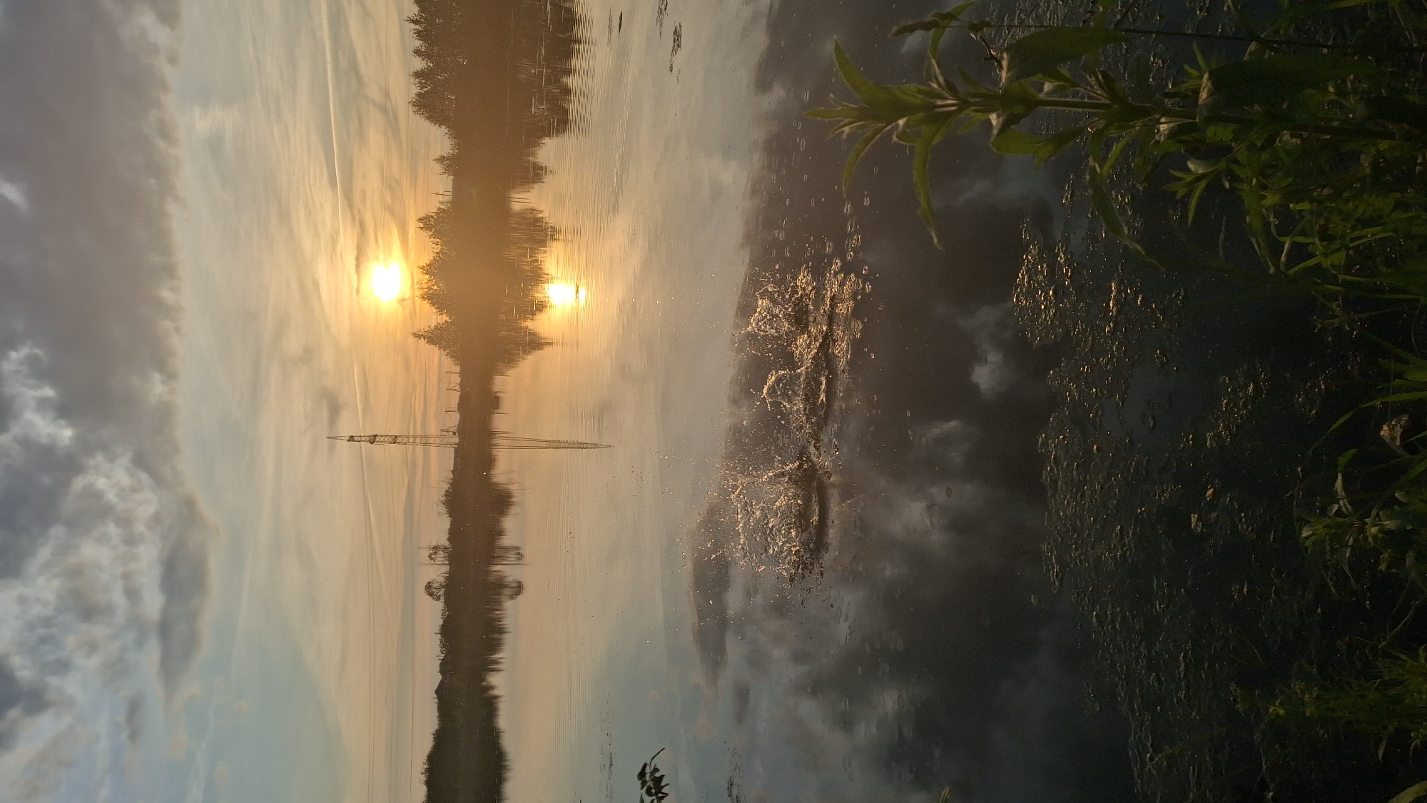 In a still body of water in Innolovo, Russia, at sunset, a splash erupts, disrupting the glassy surface. The sky is a gradient of soft blues, oranges, and grays, with scattered clouds reflecting on the water. A tall electricity pylon stands silhouetted against the horizon. Trees and buildings are faintly visible on the distant shore, their reflections mirroring in the water. The foreground is dominated by dark, murky water with patches of green algae. Green foliage grows along the water's edge, some reaching into the frame.