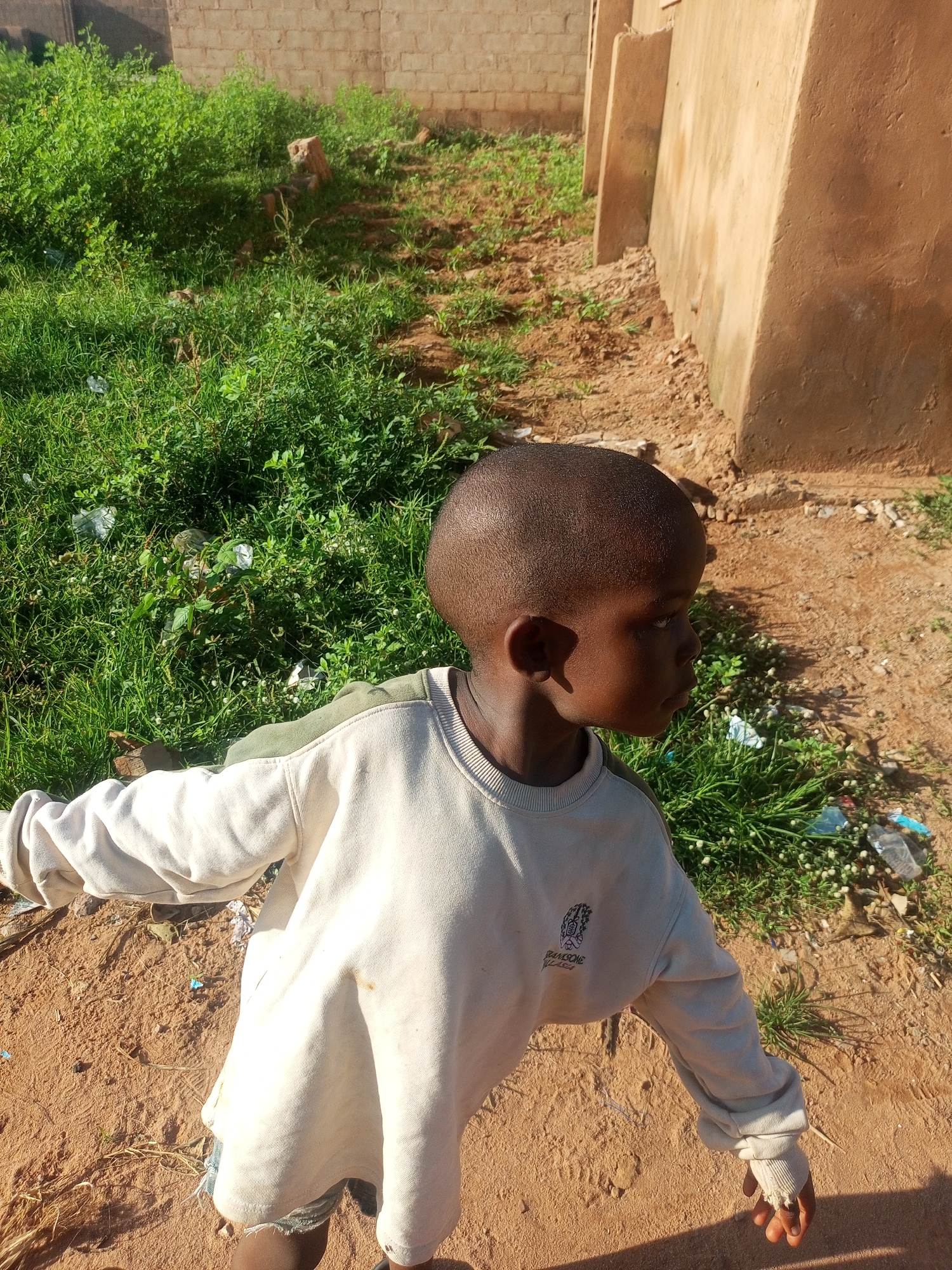 A young boy, appearing to be around five years old, stands in a dirt area with green grass and scattered debris. He is wearing a light-colored sweatshirt with some sort of emblem on the left chest and appears to be wearing ripped denim shorts. The boy is facing to his right, looking off-camera. Behind him, there is a wall constructed of light-colored bricks and a tan, rough-textured wall. The ground is sandy and uneven with patches of grass growing through. The scene appears to be outdoors, possibly in a residential or undeveloped area in Tempure, Nigeria.