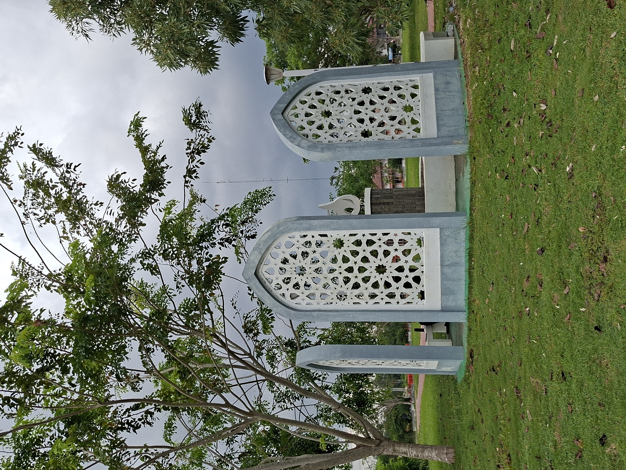 The image shows a park in Meunasahaluedrienlhoksukon Barat, Indonesia. Three decorative, arched, white structures with geometric cutouts stand on a grassy area. The structures are framed with a light grey concrete. A tree with green foliage is visible to the left of the structures. The sky is overcast with dark clouds. In the background, there are more trees and possibly buildings. Some leaves are scattered on the grass. A tall antenna or tower is visible between the first and second structure.