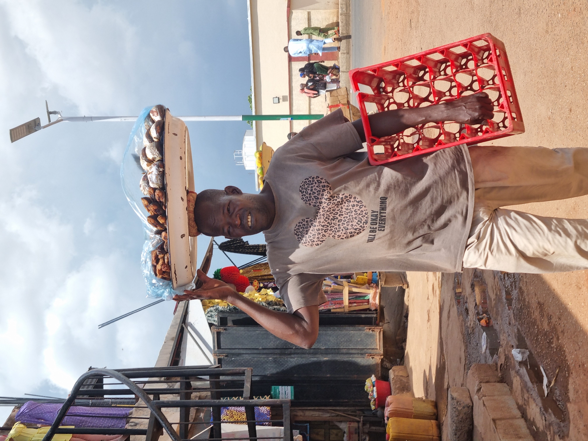 A street vendor stands on a red dirt road in Jos, Nigeria, holding a red plastic egg crate in his right hand and a tray of wrapped food on his head. The vendor has dark skin, a short haircut, and is wearing a gray t-shirt with an image of a mouse and the words "I'll Be Okay Everything" printed on it, along with tan pants. The scene is lit by bright sunlight under a sky with scattered clouds. In the background, a single light pole is erected, and people can be seen walking on a sidewalk next to a light-colored building. Below the vendor, a metal cage-like structure is visible, along with a variety of items on display for sale, including yellow and red containers. Litter is scattered on the road next to the vendor.