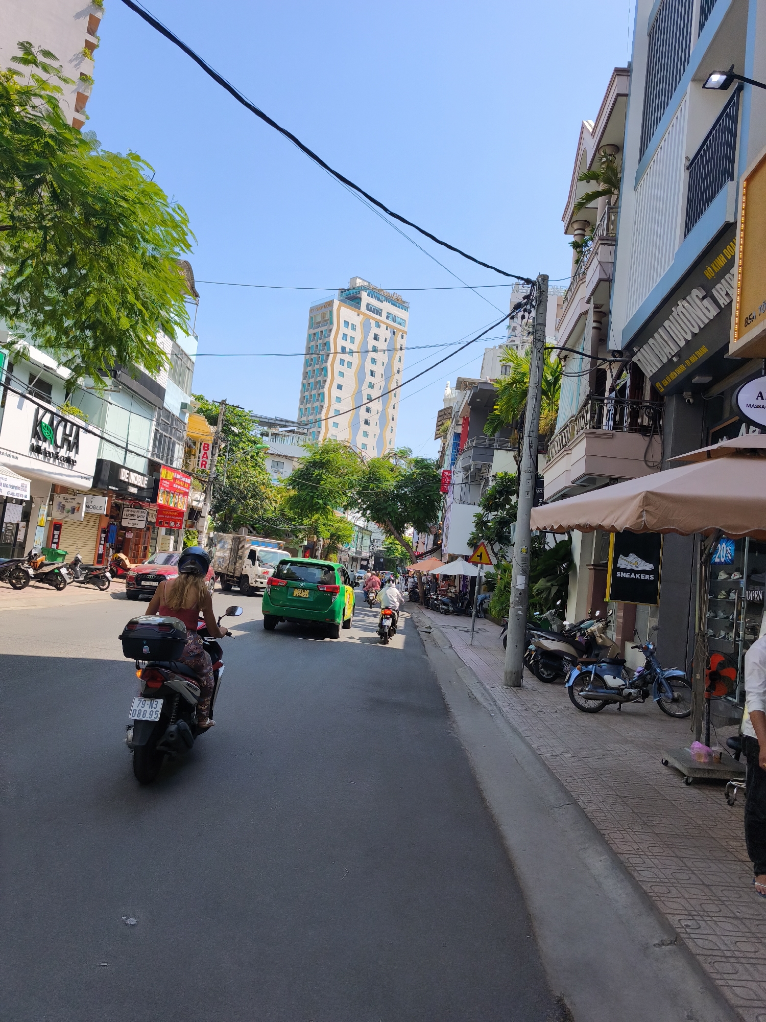The image captures a street scene in Nha Trang Ward, Vietnam, on a clear day. A woman rides a scooter down the middle of a paved road. She is wearing a red top and patterned leggings. The scooter has a black storage box on the back and a license plate that reads "79-N3 088.95". In front of her, a green car drives in the same direction. To the left of the road are several buildings, some with green trees in front. Signs on the buildings include "KACHA", "DE NOBLE", and "BAR". To the right of the road, more buildings line the street, with awnings extending over the sidewalk. A blue scooter and a beige scooter are parked on the sidewalk. One building has a sign that says "SNEAKERS" with a shoe logo. Further down the street, a tall, modern building with blue and white accents is visible. The sky is clear blue, and the sun is shining, casting shadows on the road and sidewalk.