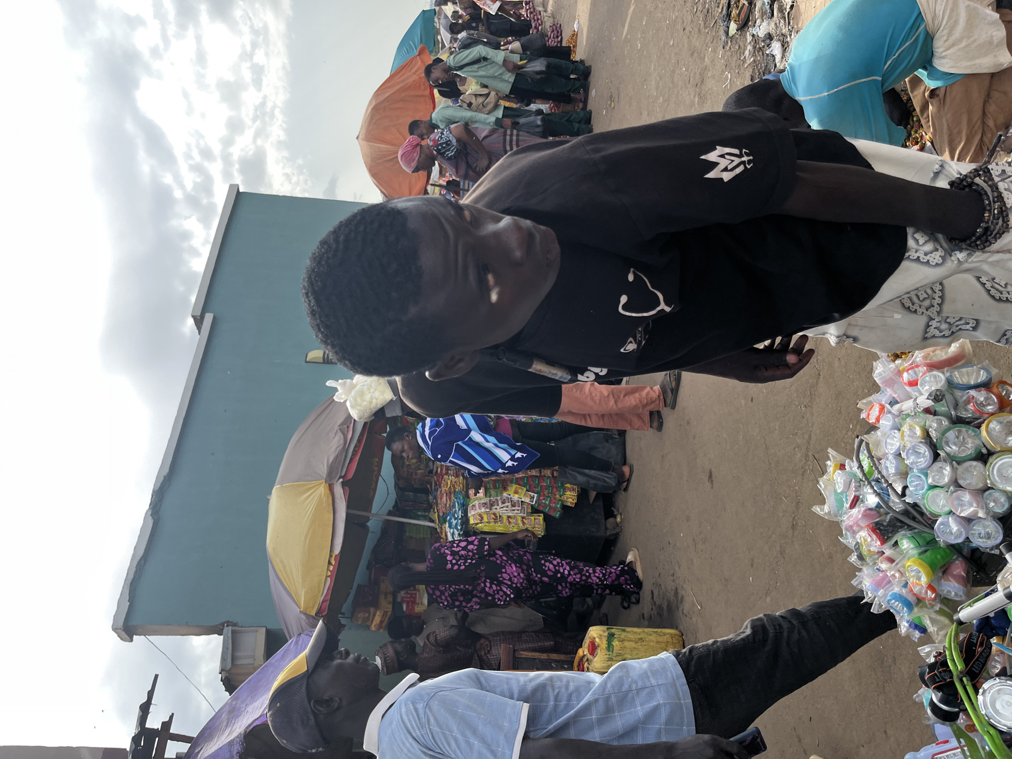 This image captures a bustling market scene in Rafin Par, Nigeria. In the foreground, a young person wearing a black t-shirt and patterned trousers looks to the right, their gaze directed downward. Behind them, a man in a blue collared shirt and dark pants stands to the left. The background is filled with activity: market stalls with colorful umbrellas, various people browsing and interacting, and piles of goods. A pile of brightly colored flashlights is visible in the bottom left of the frame. The environment appears to be an outdoor market with a dusty ground and a partially visible building in the background. The sky is cloudy, suggesting an overcast day.
