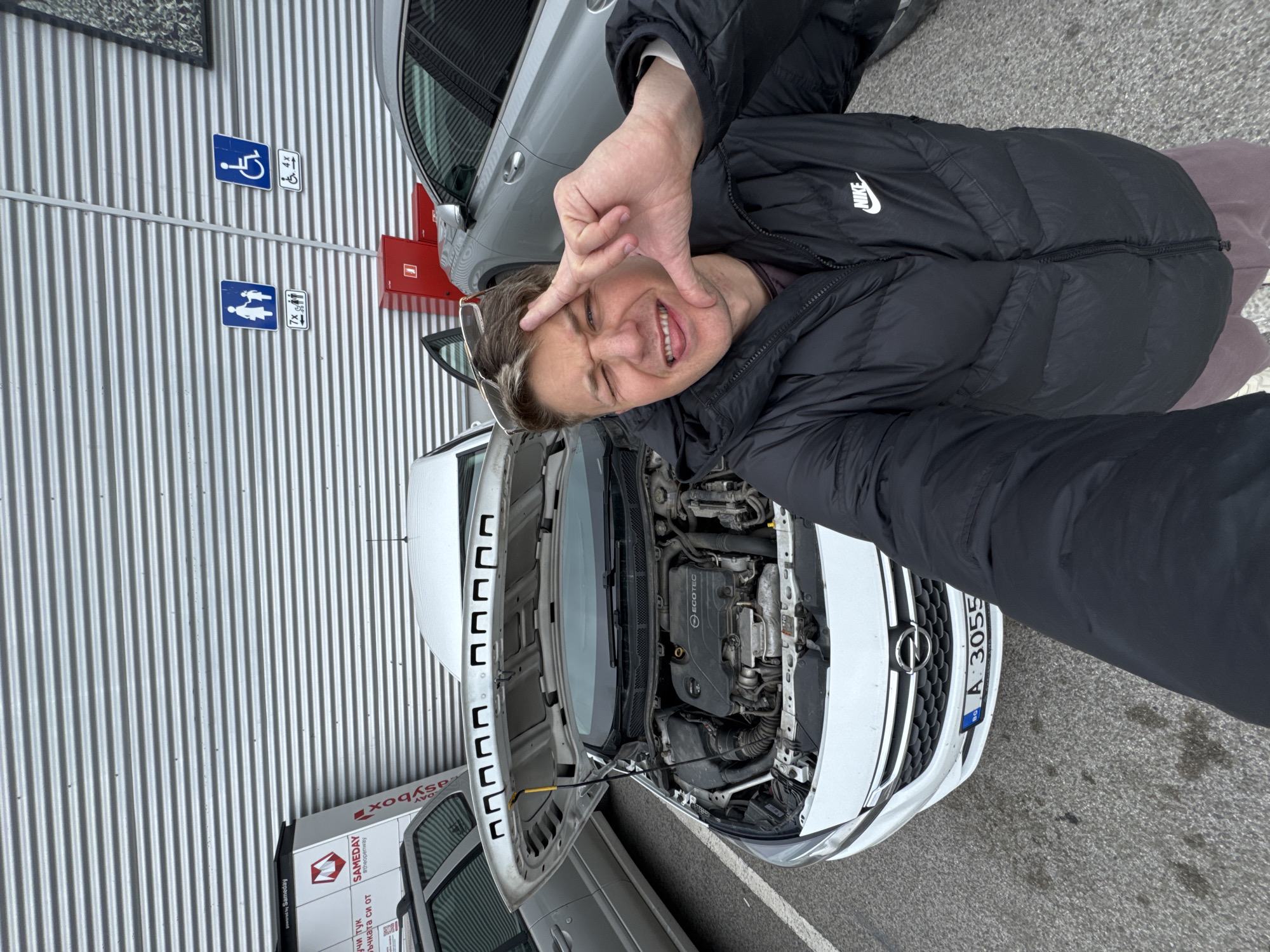 Here's a detailed description of the image:
A man wearing a black Nike puffer jacket is taking a selfie. He is smiling and making a square with his fingers near his face. Behind him is a white Opel car with its hood open, revealing the engine. Next to the Opel is a grey car, and the background shows a corrugated metal wall with blue signs indicating handicapped parking and parent-child parking spaces. The car's license plate reads "A 3055", indicating that the location is Burgas, Bulgaria.