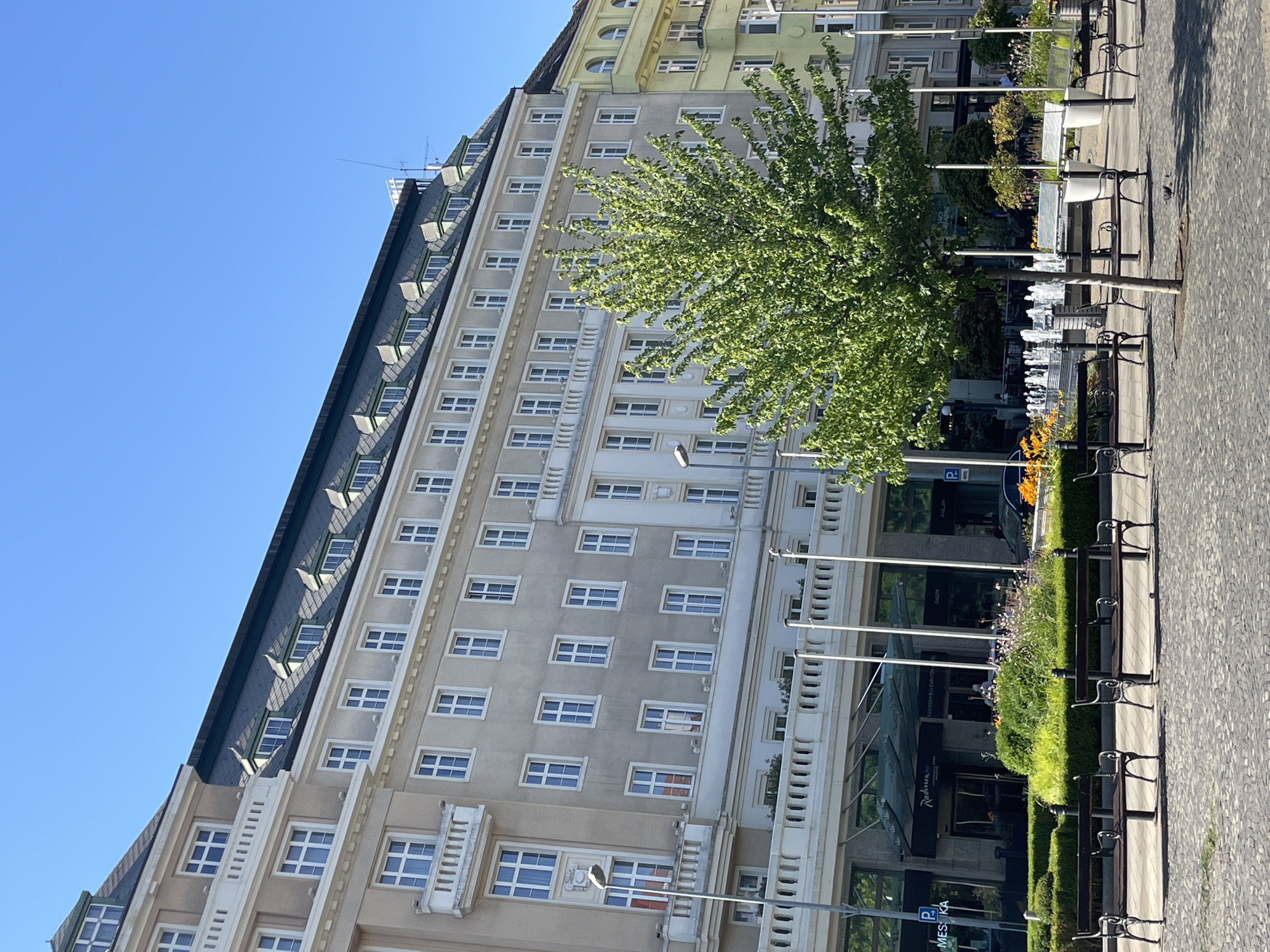 The image depicts a street scene in Bratislava, Slovakia, likely in the vicinity of Hviezdoslav Square. A large, multi-story building with a mansard roof and numerous windows dominates the background. The building appears to be a hotel, with a Radisson Blu Carlton Hotel sign visible in the lower left portion of the image. In front of the hotel, there is a pedestrian area with benches, a small tree, and a fountain. The ground is paved with cobblestones. The sky is clear and blue, indicating a sunny day. There are no people clearly visible in the frame, although there might be some figures inside the hotel entrance or very distant figures on the street. The scene is orderly and appears to be a public space in a European city.