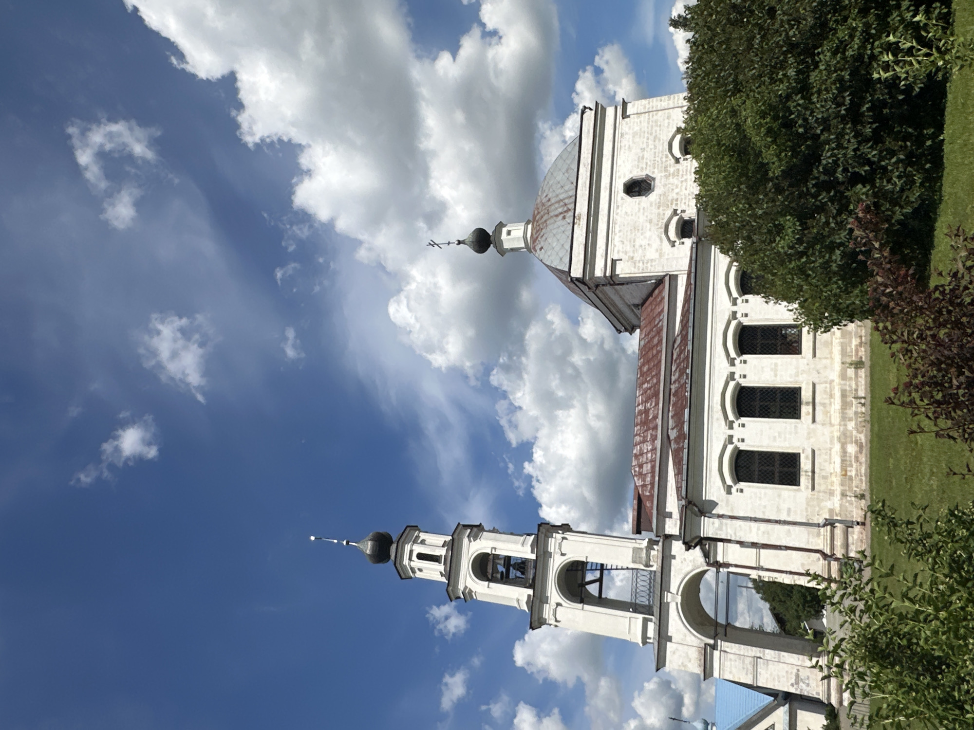 The image captures a religious structure, likely a church or cathedral, against a backdrop of a bright blue sky filled with fluffy white clouds. The architecture is predominantly white with distinct domes and bell towers, characteristic of Russian Orthodox churches. One bell tower, adorned with arches and bells, stands prominently, topped by a dark, onion-shaped dome and a cross. Adjacent to it, a larger dome with a similar cross rises from the main building. The building exhibits multiple windows with arched tops and a reddish-brown roof. Lush green grass surrounds the structure, with dense trees and bushes framing the right side of the image.
The lighting suggests it is daytime, likely midday or early afternoon, given the bright sky and the way light falls on the building. The weather appears to be pleasant, with a mix of sun and clouds. The overall scene evokes a sense of tranquility and architectural grandeur. No people are visible in the frame. The location is identified as Erino, Russia. No text is visible in the image.