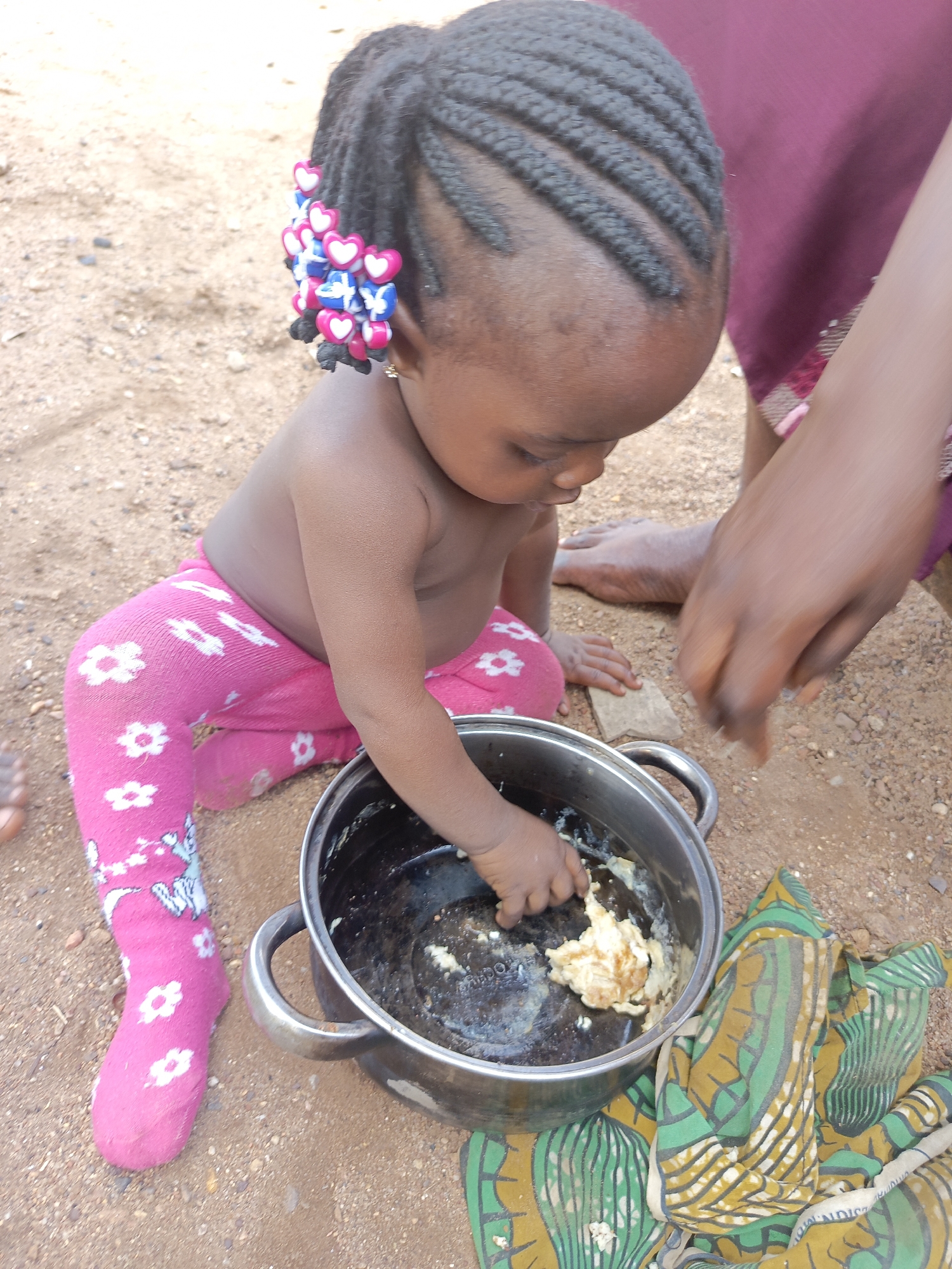 In Akwanga, Nigeria, a young child with braided hair adorned with heart-shaped beads sits outdoors on a dirt surface, wearing pink pants with white flower patterns. The child reaches into a silver pot containing what appears to be cooked food residue. A nearby adult hand offers assistance, while a patterned cloth rests beside the pot on the ground.