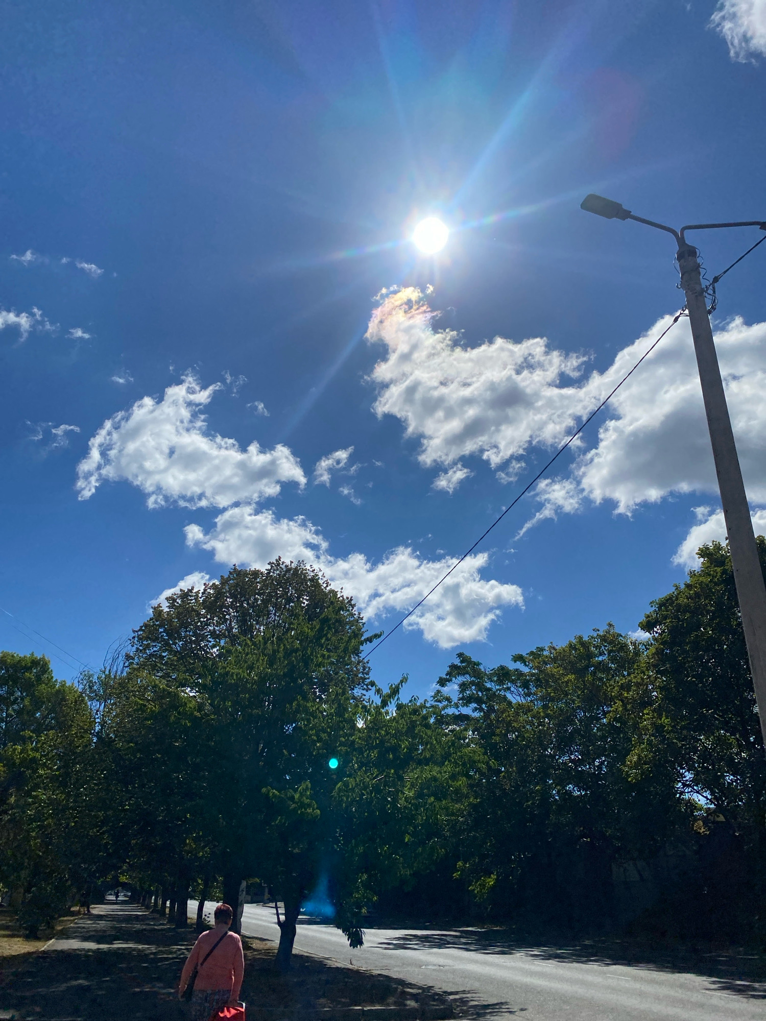 The image depicts a street scene in Chornomorsk, Ukraine, under a bright, sunny sky with scattered white clouds. The sun is positioned high in the sky, creating lens flare and casting a bright glow. A street lamp with a grey pole stands on the right side of the frame, with electrical wires stretching across. Dense green trees line the left side of the asphalt road, creating a shaded walkway. A person wearing a pink shirt and patterned pants is walking away from the camera on the walkway, carrying a red bag and a shoulder bag. The road is paved and appears to be a public street. The overall environment suggests a daytime scene in a town or city.