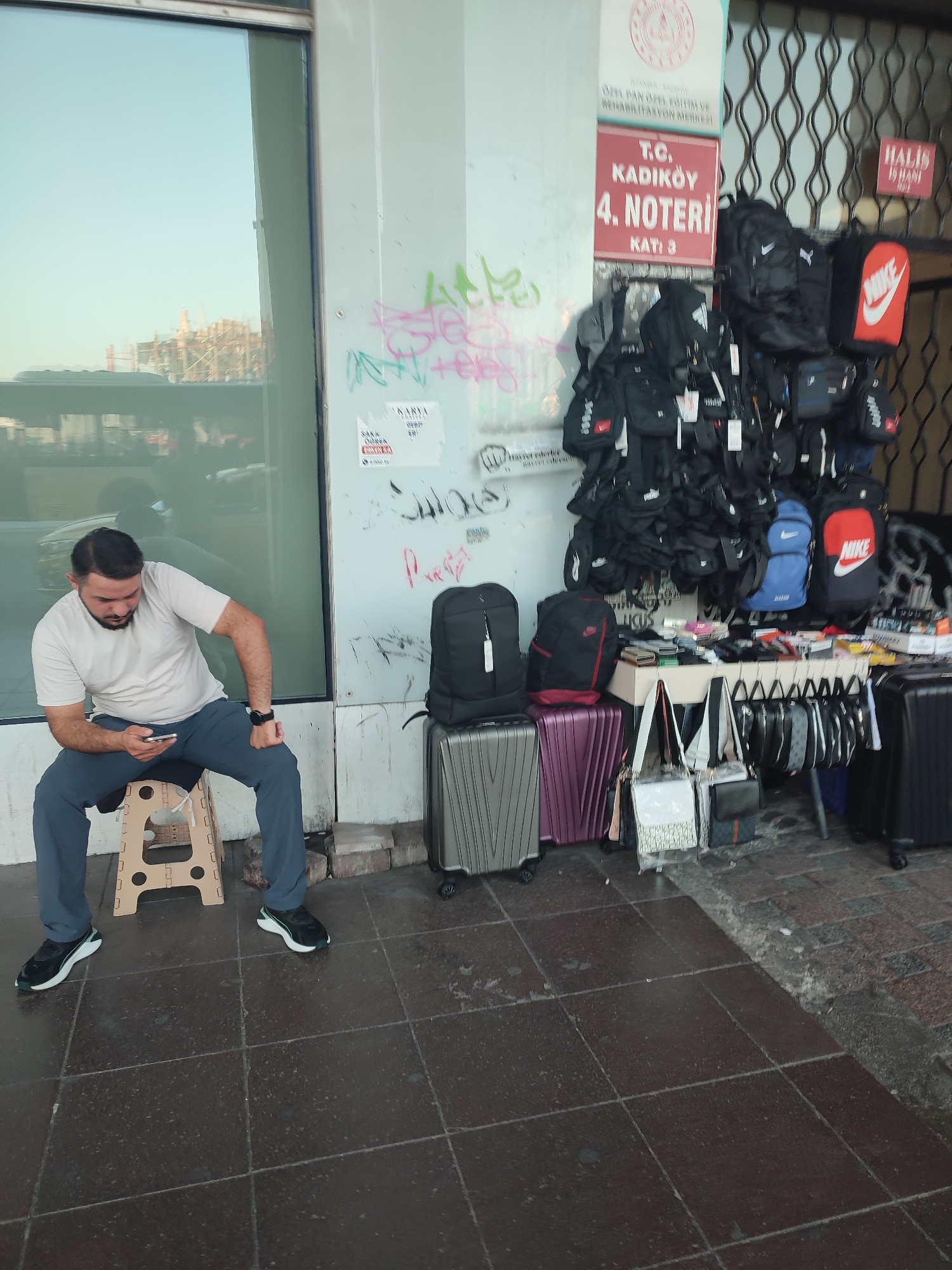 A man is seated on a beige, folding stool outdoors, looking at his smartphone. He wears a light-colored t-shirt and dark pants, with athletic shoes. To his right, there are several suitcases, including one silver, one purple, and one black. Behind him, a display rack holds numerous black bags, primarily backpacks and crossbody bags, many with brand logos such as Nike and Puma visible. A red sign with white text that reads "T.C. KADIKÖY 4. NOTERİ KATI 3" is affixed to a white wall. This indicates the location is the 4th notary office in Kadıköy, Turkey, on the third floor. Graffiti is present on the wall to the left of the sign. The ground is paved with dark, square tiles.