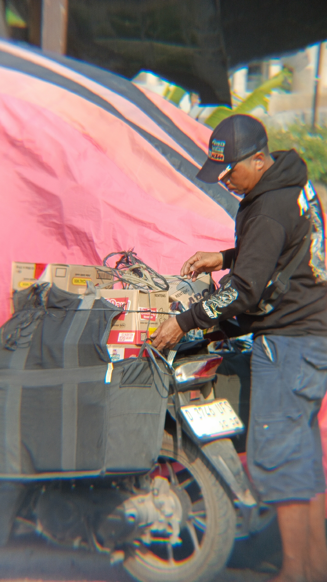 In Margajaya, Indonesia, a person wearing a black baseball cap and a black hoodie with graphics on the sleeves is standing next to a motorcycle. The motorcycle is loaded with several cardboard boxes, some of which are labeled "RENTENG" and "ABC." A large grey insulated bag is strapped to the back of the motorcycle, over the boxes. The person is reaching out with their right hand towards the boxes, appearing to adjust or secure them. The background consists of a large, taut pink and black fabric, possibly a canopy or banner, and some blurred greenery. The lighting suggests it is daytime. The license plate on the motorcycle is partially visible.