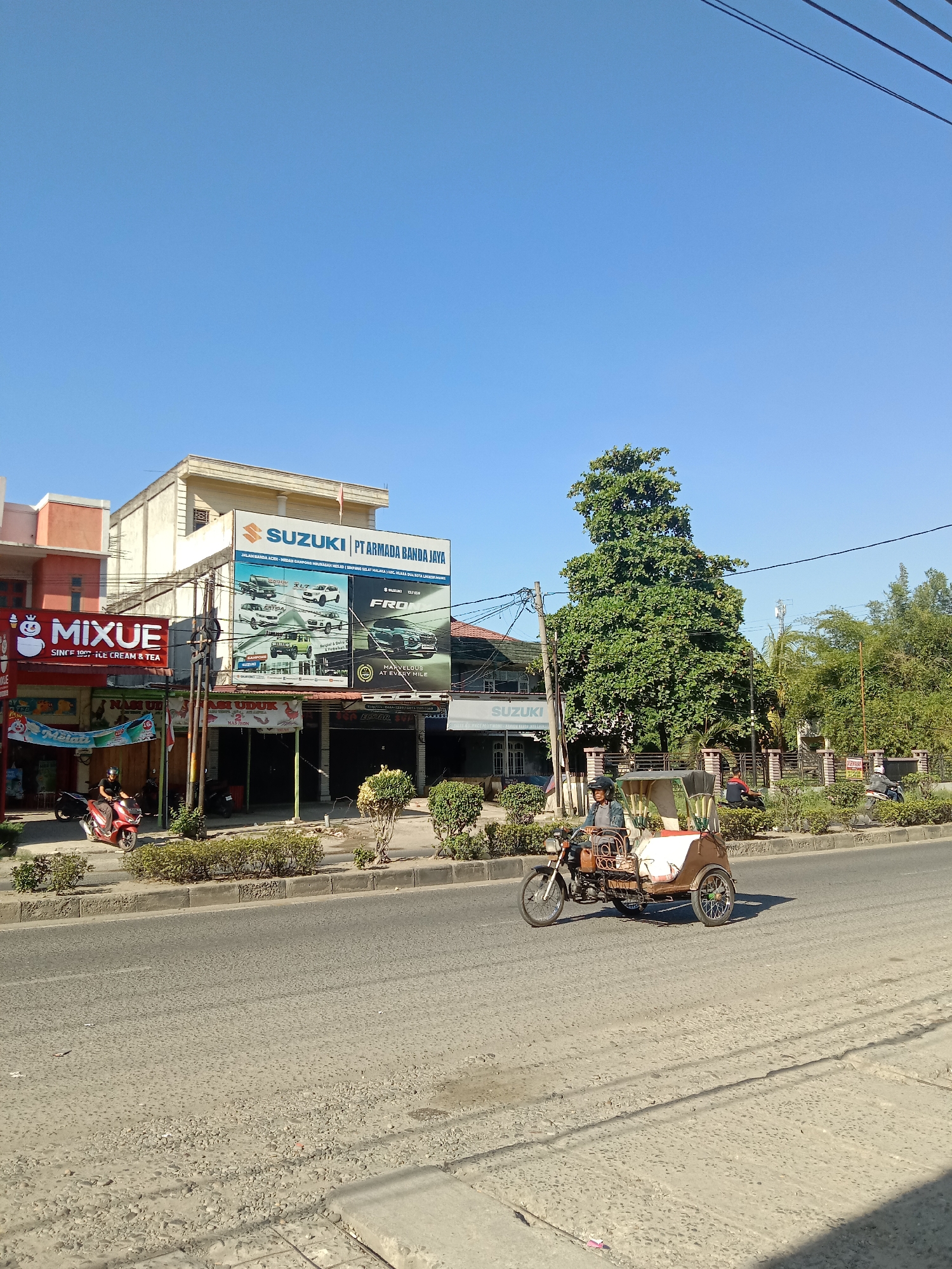 The image captures a street scene in Lhokseumawe, Indonesia, under a clear blue sky. Prominently featured on the left is a two-story building housing a "MIXUE" ice cream and tea shop, identified by its red and white signage. Adjacent to it is a Suzuki dealership, "PT ARMADA BANDA JAYA", with a large billboard displaying various Suzuki vehicle models. The road in the foreground shows signs of wear and tear. A motor tricycle, or "becak," is actively traveling down the road, manned by a driver. A few other motorbikes are parked in front of the shops. Street-side landscaping with small bushes is visible between the road and buildings. Across the street, a fenced property with trees adds depth to the background.
