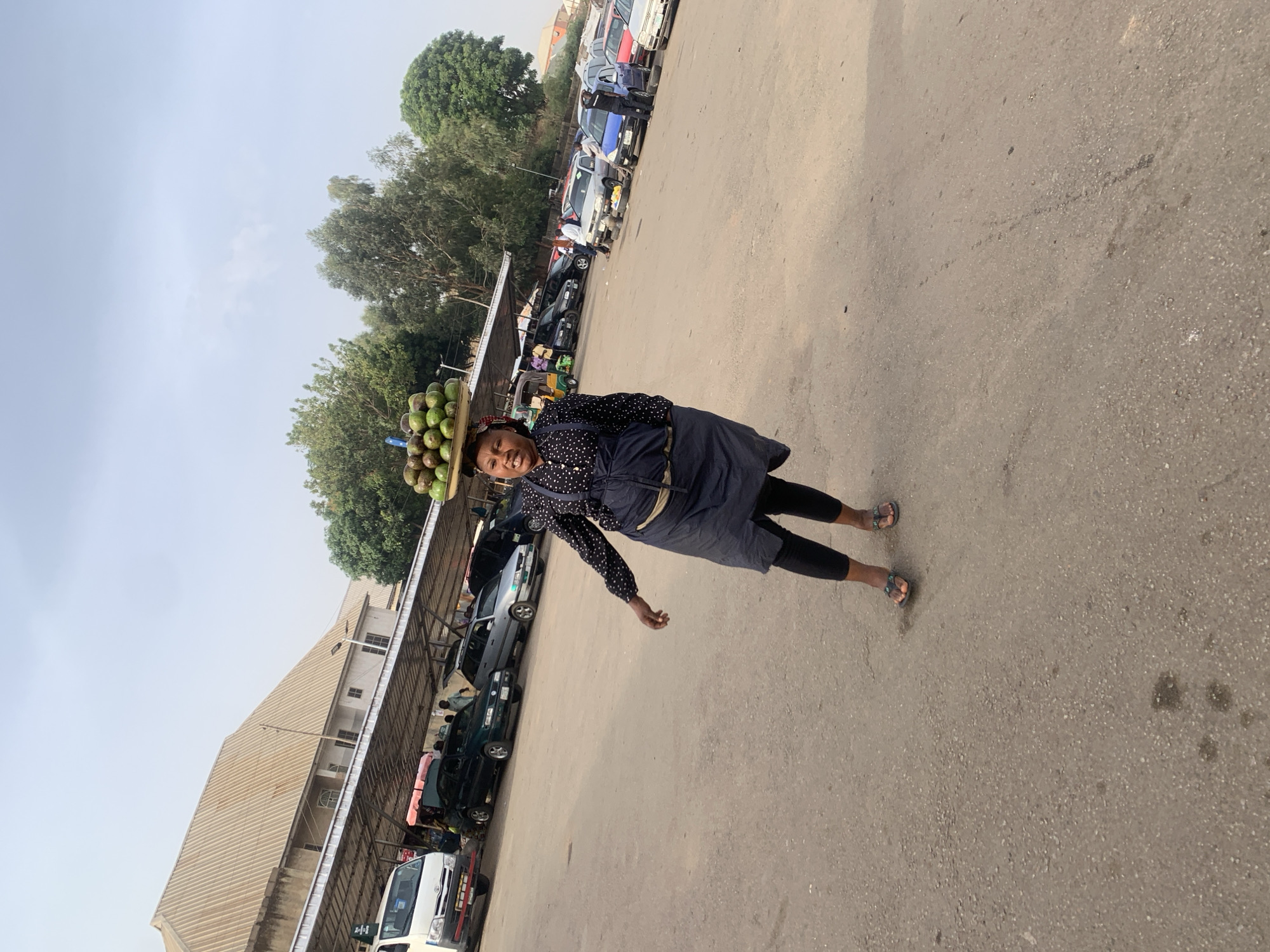 The image depicts a woman in Bukuru, Nigeria, carrying a tray of green coconuts on her head. She wears a polka-dot shirt, dark skirt, and sandals. She is walking on a paved surface, possibly a road or parking lot, lined with parked cars and a building with a corrugated metal roof. The sky is overcast.
