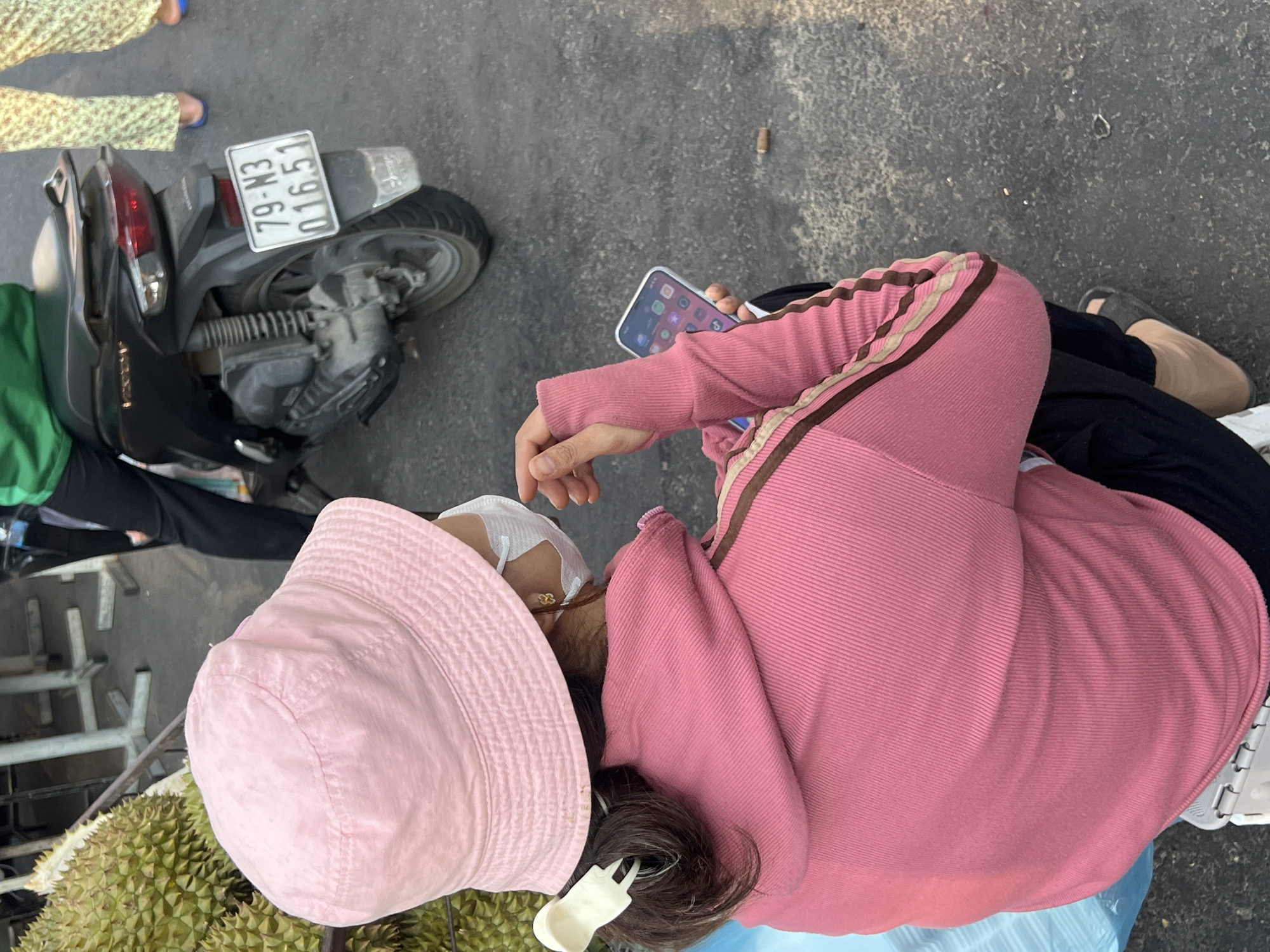 A high-angle shot shows a woman wearing a pink bucket hat, a white face mask, and a pink jacket with brown stripes on the sleeves. She's holding a smartphone and is seated near a durian fruit. The woman's hair is dark and pulled back with a white clip. In the background, a motorcycle with the license plate "79-N3 016.51" is visible. The scene appears to be a street, and based on the license plate, it's likely in Nha Trang, Vietnam.