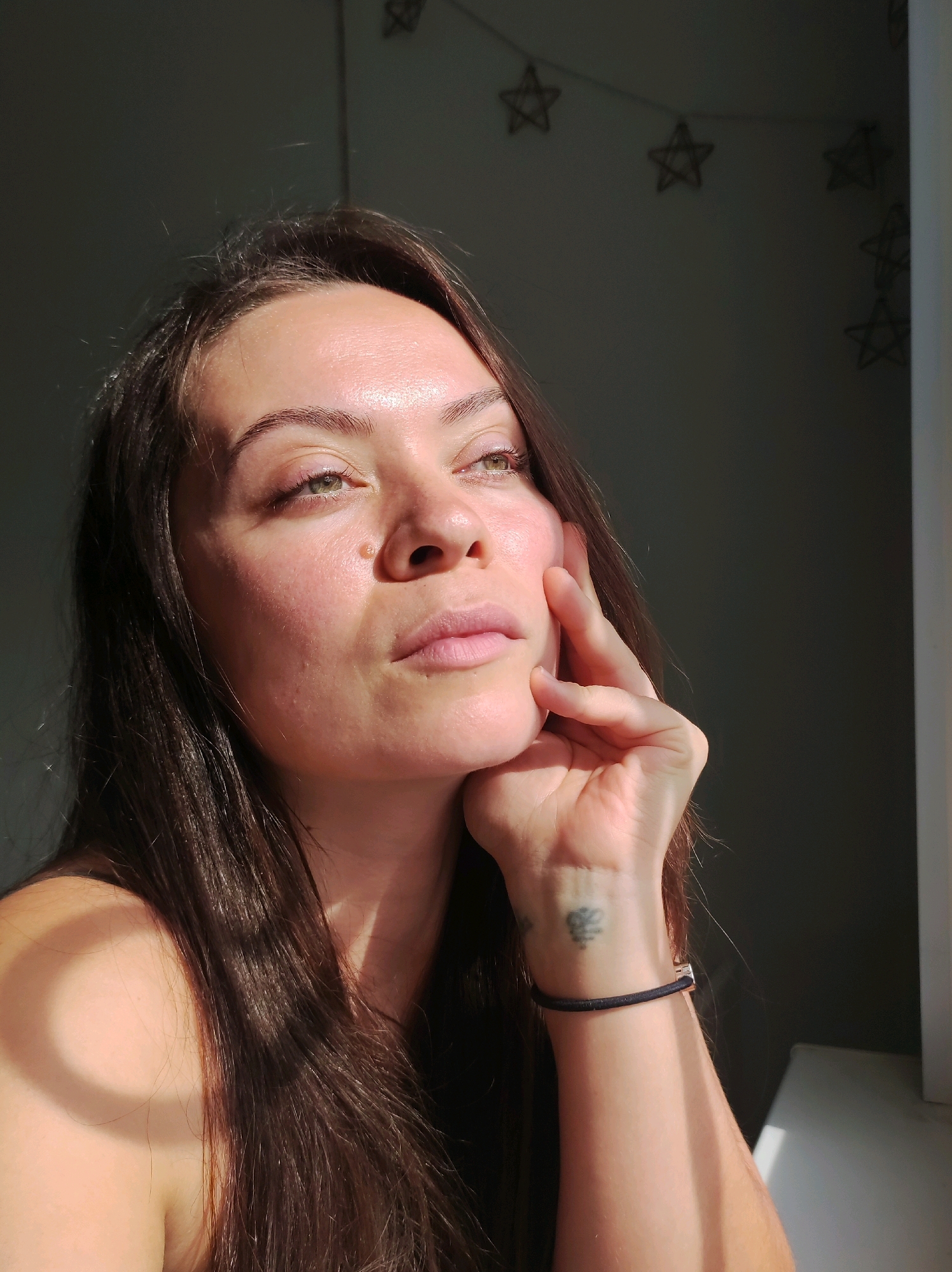 A medium close-up shot shows a young woman with long, dark brown hair looking up and to her right, with her left hand resting on her cheek and chin. The woman has light skin with some redness and slight imperfections, and her eyes are open and appear to be green. She is looking slightly off-camera, with a pensive expression. The lighting suggests natural light coming from the right, creating highlights on her face and shoulder, and casting shadows on the left side of her face and on the wall behind her. There are string lights shaped like stars visible in the background, hanging on a dark wall. A portion of a white windowsill is visible on the right side of the frame. The overall impression is one of quiet contemplation in a domestic setting. No specific location in Chaikovsky, Russia can be identified from the image.