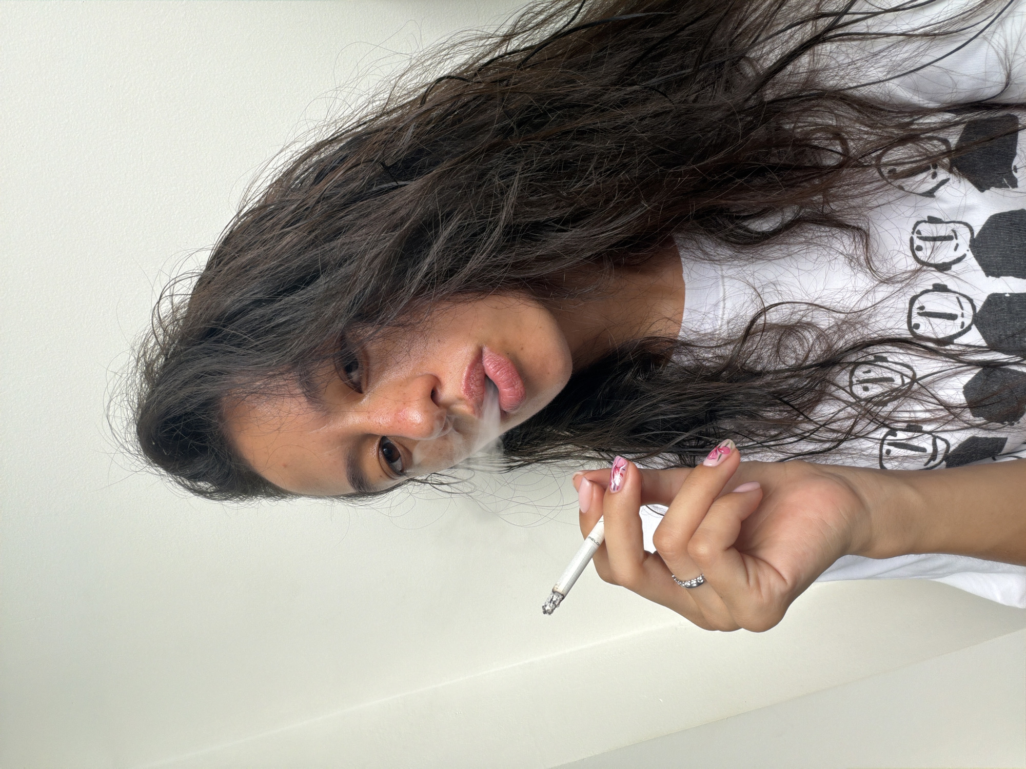 A young woman with long, dark, wavy hair is shown from the chest up, looking directly at the camera and exhaling smoke from a cigarette. She is wearing a white t-shirt with a black graphic print of multiple stylized faces. Her right hand, holding the cigarette, is positioned near her face, with a silver ring on her left ring finger and her nails painted with a pink and white floral design. The background is a plain, light-colored wall, and the lighting appears to be natural, coming from the left. The image is a close-up selfie, with the camera positioned slightly above and to the left of the subject.
