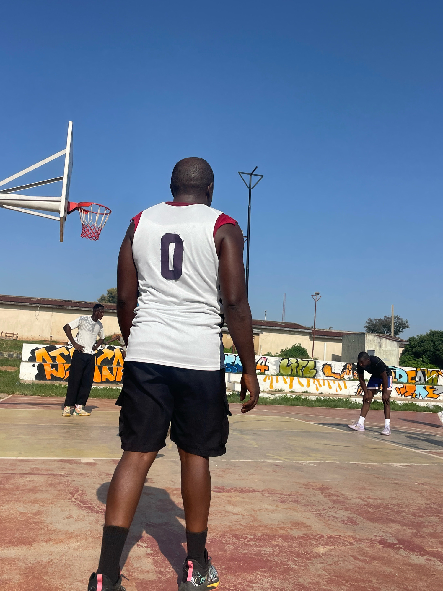 In an outdoor basketball court in Jos, Nigeria, three individuals are present. The primary subject is a man standing in the foreground, facing away from the camera, wearing a white basketball jersey with the number "0" on the back, black shorts, and black socks. To his left, another man stands, also facing away, wearing a white t-shirt and dark pants. In the background, a third man is seen bending down, wearing a dark shirt and blue shorts. The court surface is visible, marked with lines, and a basketball hoop with a red net is mounted on a white backboard. A wall with colorful graffiti runs along the perimeter of the court, behind the players. The sky is clear and blue.