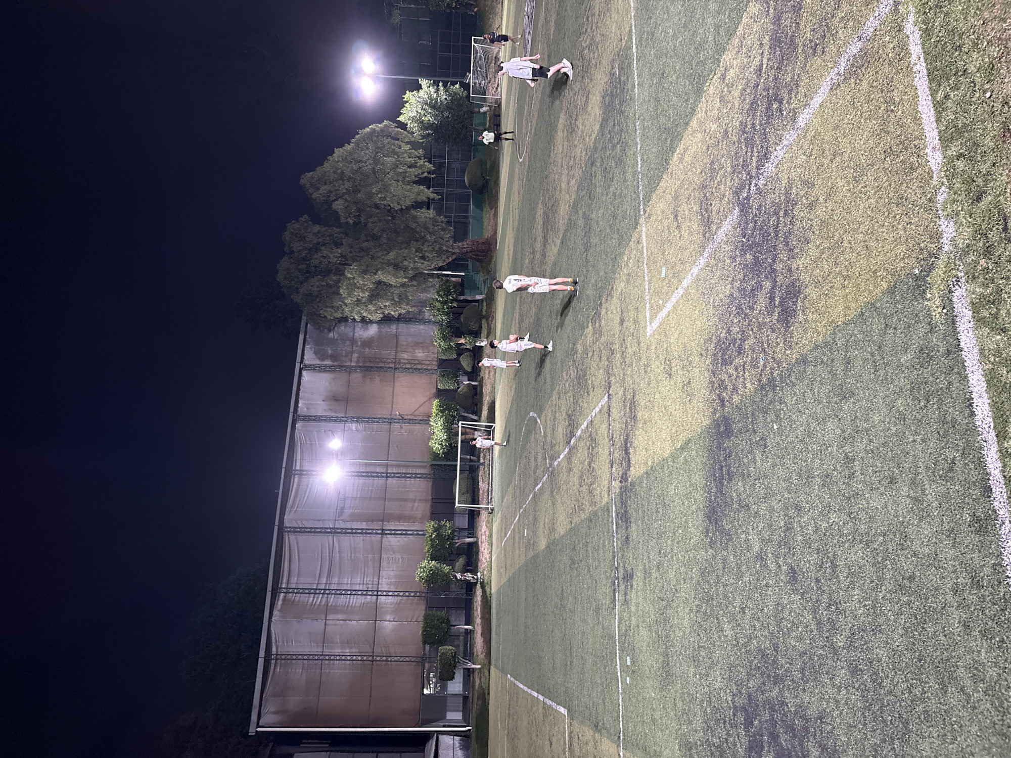 The image depicts an artificial turf soccer field illuminated by stadium lights at night. Several people are dispersed across the field. In the background, a structure appears to be a building or enclosed area adjacent to the field, also illuminated by artificial lights. Trees are scattered around the perimeter of the field. The image was likely taken in Huixquilucan de Degollado, Mexico.