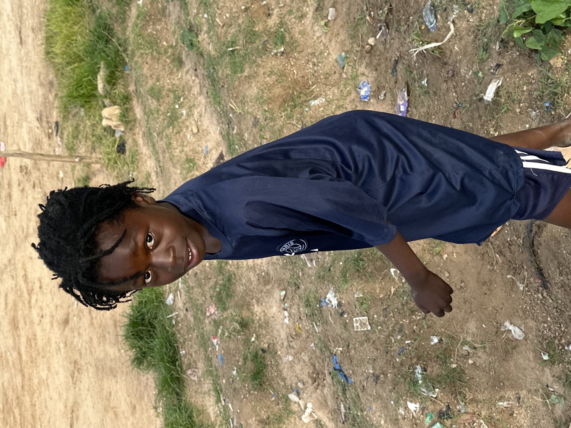 The image shows a young Black person, likely a child, with dark dreadlocks and brown eyes, looking towards the camera. They are wearing a dark blue t-shirt with a logo that appears to say "PARIS" and some smaller text underneath, and dark blue shorts. The person is standing outdoors on uneven, dry ground with patches of sparse green grass. The ground is littered with various pieces of debris, including plastic wrappers and other refuse, suggesting an unkempt environment. In the background, there are hints of further greenery and more scattered trash. The lighting suggests it is daytime, possibly late morning or afternoon, with no strong shadows. The person's expression is friendly and open, with a slight smile. The overall scene appears to be a casual, everyday setting in a rural or semi-rural area, potentially in Nigeria given the location context provided. The image is oriented with the subject's head towards the left side of the frame, suggesting it was taken in portrait mode and then rotated.