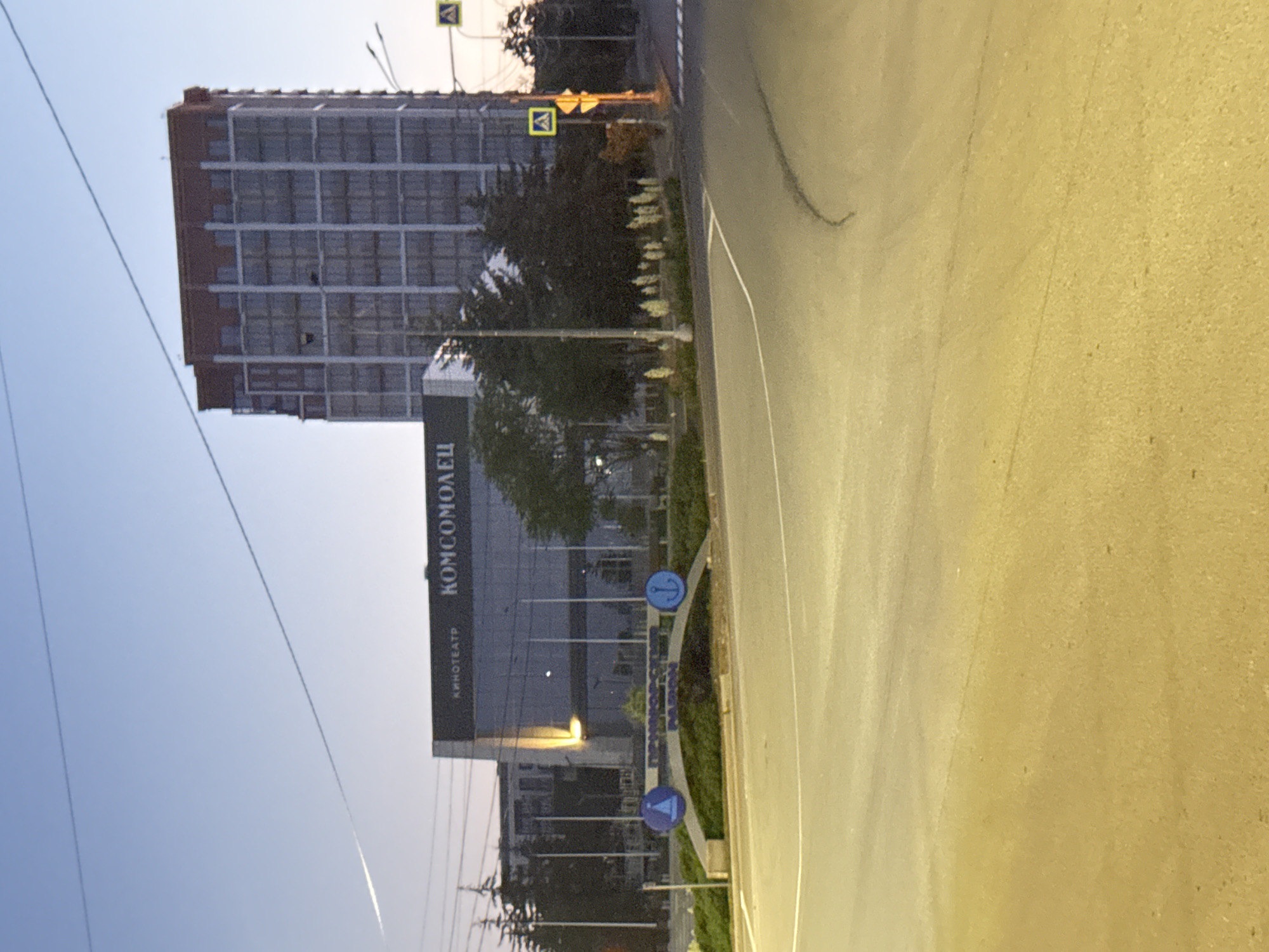 The image depicts the city of Mariupol, Ukraine, at dusk or dawn. In the background, a tall, modern building with a red-capped roof is visible, possibly an apartment complex or hotel, featuring numerous windows. In front of it, a lower, dark-colored building with large glass windows bears the inscription "КИНОТЕАТР КОМСОМОЛЕЦ" (Cinema Komsomolets), indicating a cinema complex. To the left of the cinema, a bright yellow light shines from an interior source.
In the mid-ground, a decorative archway spans across a green, landscaped area. The archway is inscribed with Cyrillic text, "ПРИМОРСКИЙ РАЙОН" (Primorsky District), and features circular blue emblems with nautical motifs – a sailing ship on the left and an anchor on the right. Several streetlights and trees are scattered around this area.
The foreground shows a paved road with white lane markings, suggesting a street intersection or roundabout. The lighting in the foreground is warm and yellow, likely from streetlights. The sky is a pale blue, with a faint white contrail from an aircraft visible. Traffic signs, including a pedestrian crossing sign, are present on the right side of the image. There are no visible people or vehicles in the scene.