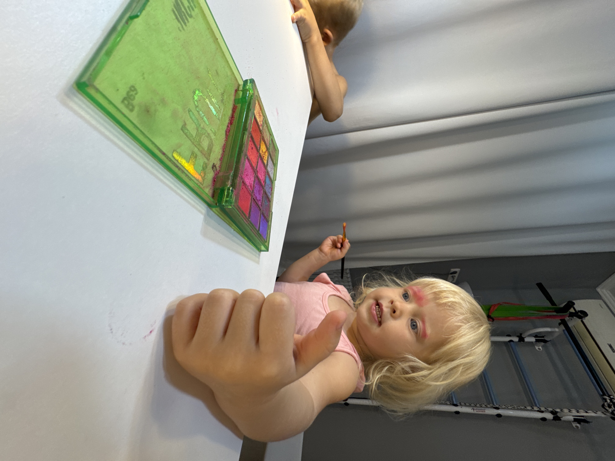 A young, blonde, fair-skinned child is captured in a medium-shot photograph. The child is situated next to a table. The child appears to be a toddler, wearing pink on their eyebrows. The child is holding a small paintbrush and interacting with a makeup palette sitting on a white table. The table is also stained with the child's makeup hand prints. There is another child with blonde hair visible behind the table. The photo was taken inside a room with white walls and dark floors. There is equipment visible in the background suggesting it could be Sochi, Russia.