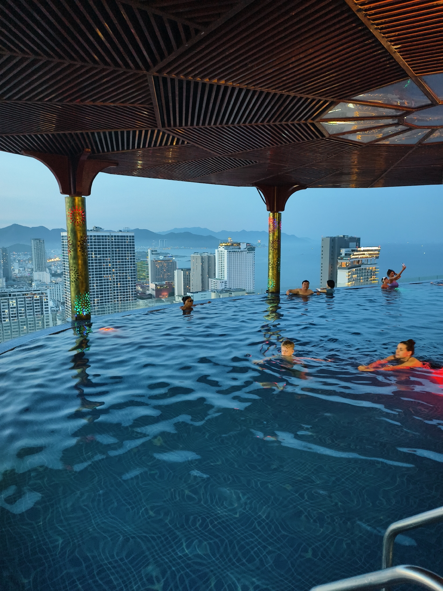 This image captures an evening scene at an infinity rooftop pool in an unidentified city. The sky, a gradient of light to deep blue, suggests dusk or early night, with silhouetted mountains visible in the distant background. A sprawling urban landscape featuring numerous illuminated high-rise buildings extends beyond the pool's edge, leading to a body of water on the right horizon.
Above the pool, a distinctive overhead structure with dark, geometrically patterned slats provides shade or architectural interest. This structure is supported by several ornate pillars; one prominent golden-colored pillar on the left features illuminated, multi-colored cut-out patterns (red, green, blue).
In the foreground, the dark blue water of the infinity pool exhibits ripples and reflects ambient light and portions of the overhead structure. Multiple individuals are present in the pool:
- On the far right, a woman in a purple float takes a selfie with a phone, accompanied by another person behind her.
- Towards the center-right, a woman with her back to the camera and a man with short dark hair face the city view, resting on the pool's edge.
- In the mid-ground, a young boy with light hair floats, looking towards the left.
- Closer to the foreground on the right, a woman is illuminated by a red light from beneath the water, looking left.
- A metal handrail for pool access is visible in the bottom right corner.