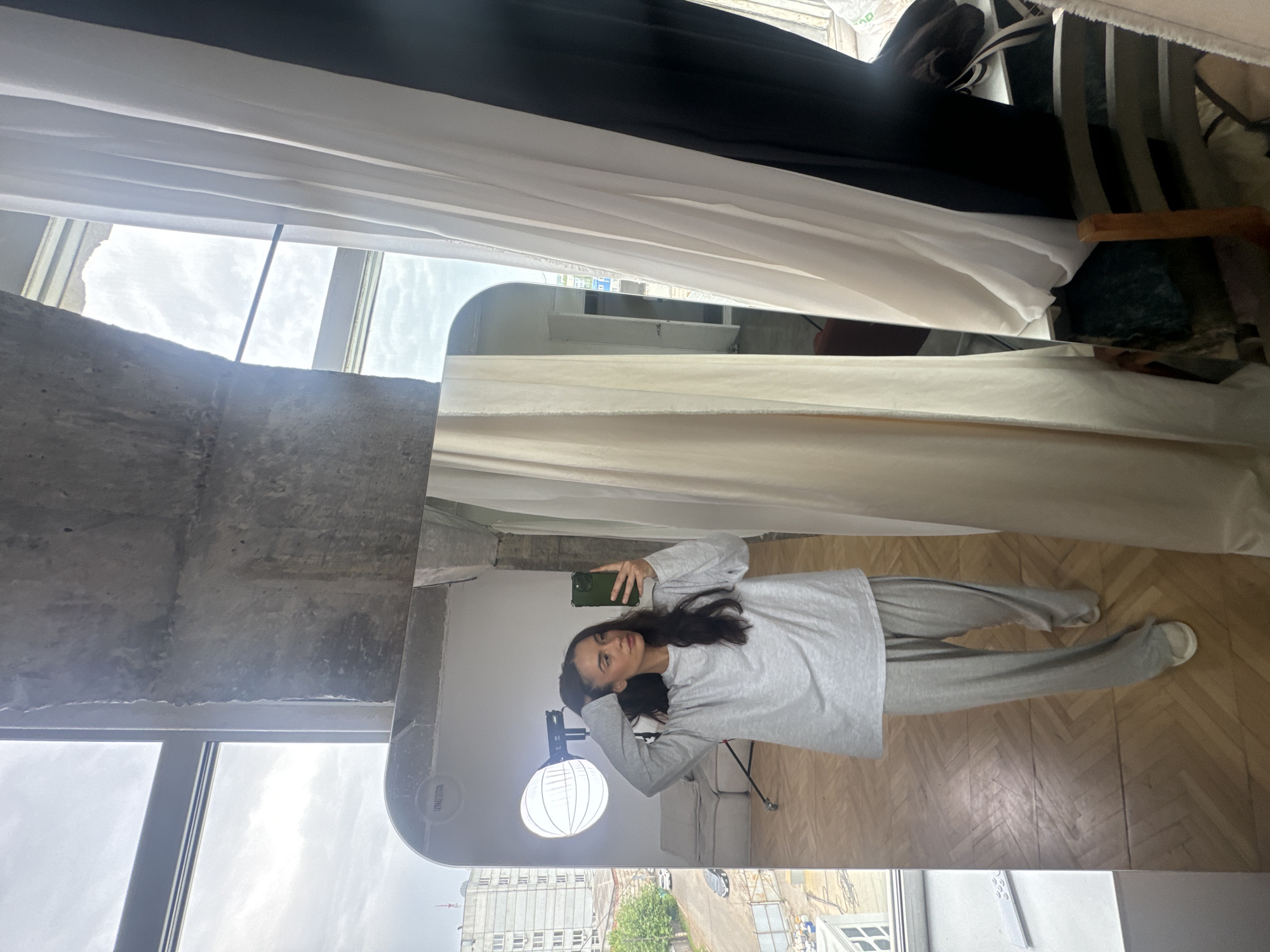 A young woman stands in front of a large, full-length mirror, taking a selfie with her smartphone. She is wearing a light gray long-sleeved t-shirt and matching loose-fitting pants, with white sneakers. Her dark hair is pulled back slightly, and she is looking at her reflection. The mirror reflects the room she is in, which appears to be a studio or loft-style apartment with large windows. Through the windows, a city skyline is visible, featuring apartment buildings and trees. One of the windows looks out onto a street with cars and a building under construction. The interior of the room has exposed concrete elements and wooden flooring. A large softbox light, commonly used in photography, is positioned behind the woman, also reflected in the mirror. The overall scene suggests a personal space, possibly a home studio or a creatively styled living area. The architectural style and the visible urban landscape indicate the location is likely Krasnoyarsk, Russia, a major city in Siberia.