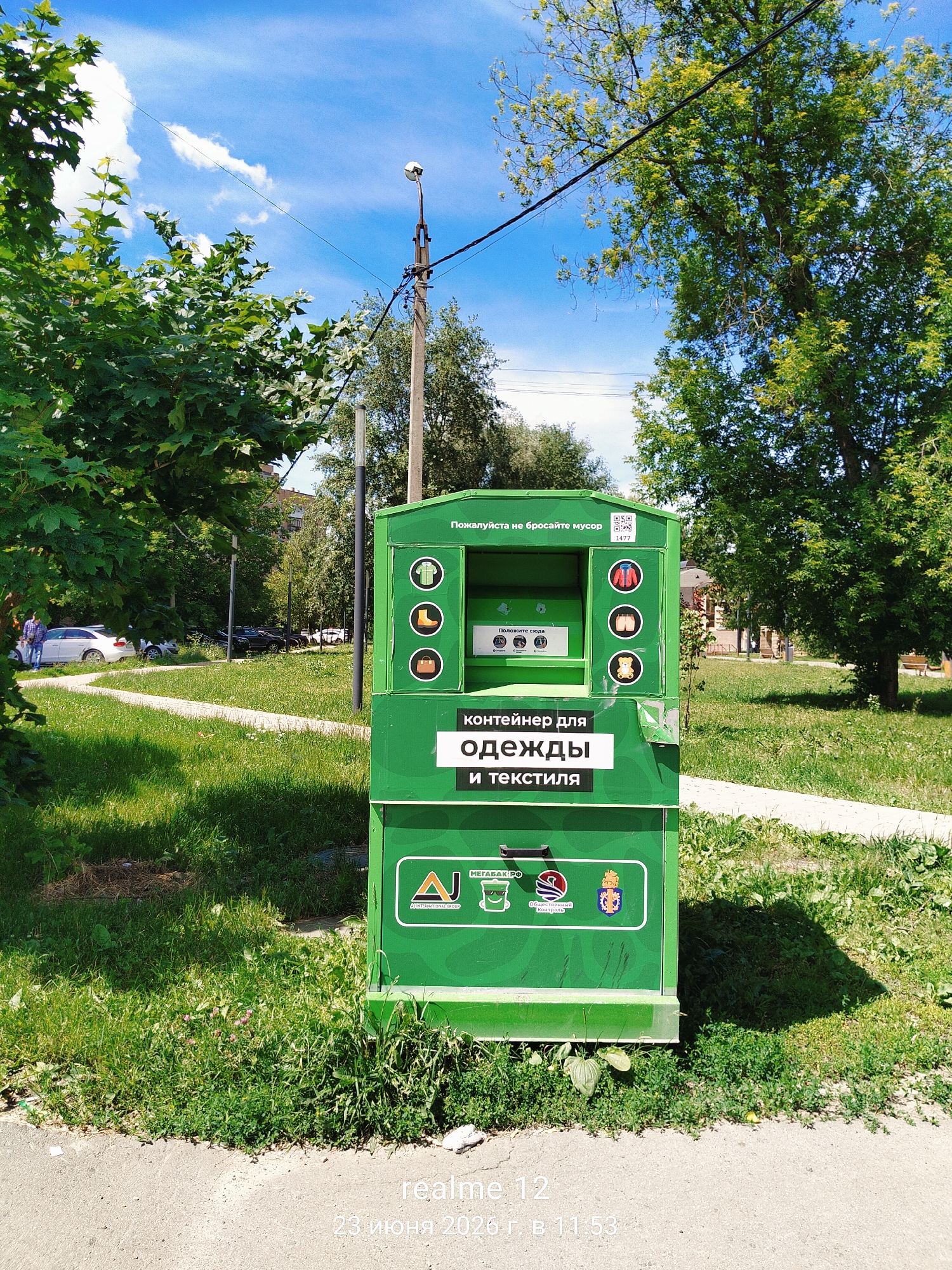An outdoor shot captures a bright green recycling container for clothes and textiles in Shchyolkovo, Russia. The container is positioned on a grassy area beside a sidewalk, with trees and a blue sky with white clouds in the background. The container has several symbols depicting clothing items, such as a t-shirt, boots, and jackets, and text in Russian that translates to "Container for clothes and textiles." A QR code and the number 1477 are also visible on the container. To the left of the container, a white car and a few people are visible in the distance. The lower right corner of the image shows text that reads "realme 12 23 июня 2020 г. в 11:53," indicating the date and time the photo was taken.