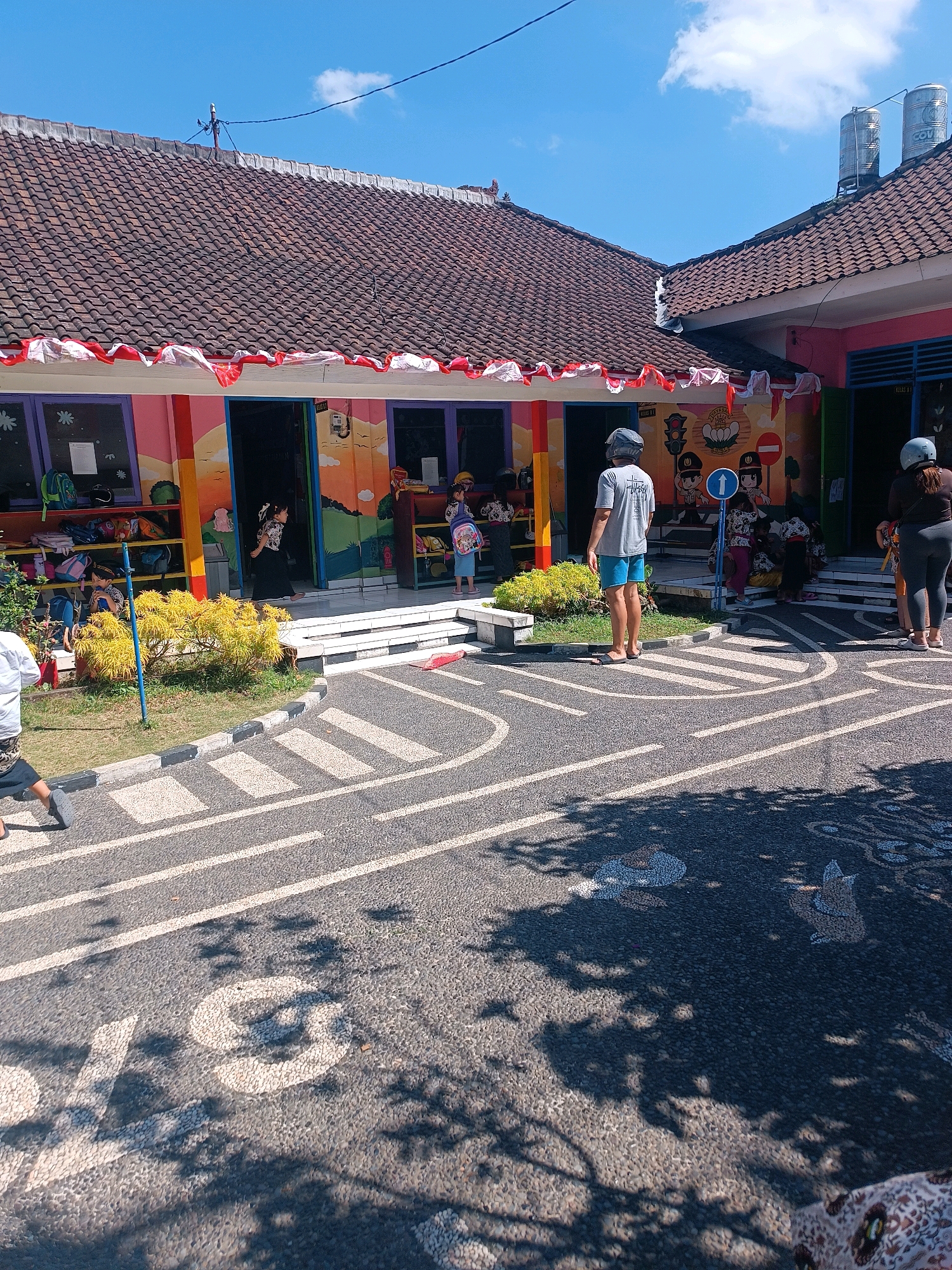 The image shows the exterior of a colorful building with red and white decorations along the roofline. The building appears to be a school or kindergarten, as indicated by the children and staff present and the playful murals on the walls. In front of the building is a simulated road with white lines and a crosswalk, suggesting a traffic safety education area for children. Several children are visible in the doorway and on the steps of the building, some wearing backpacks. Adults, some wearing helmets, are standing on the road and near the entrance. The sky is clear and blue. The context provided indicates the location is the city of Tabanan, Indonesia.