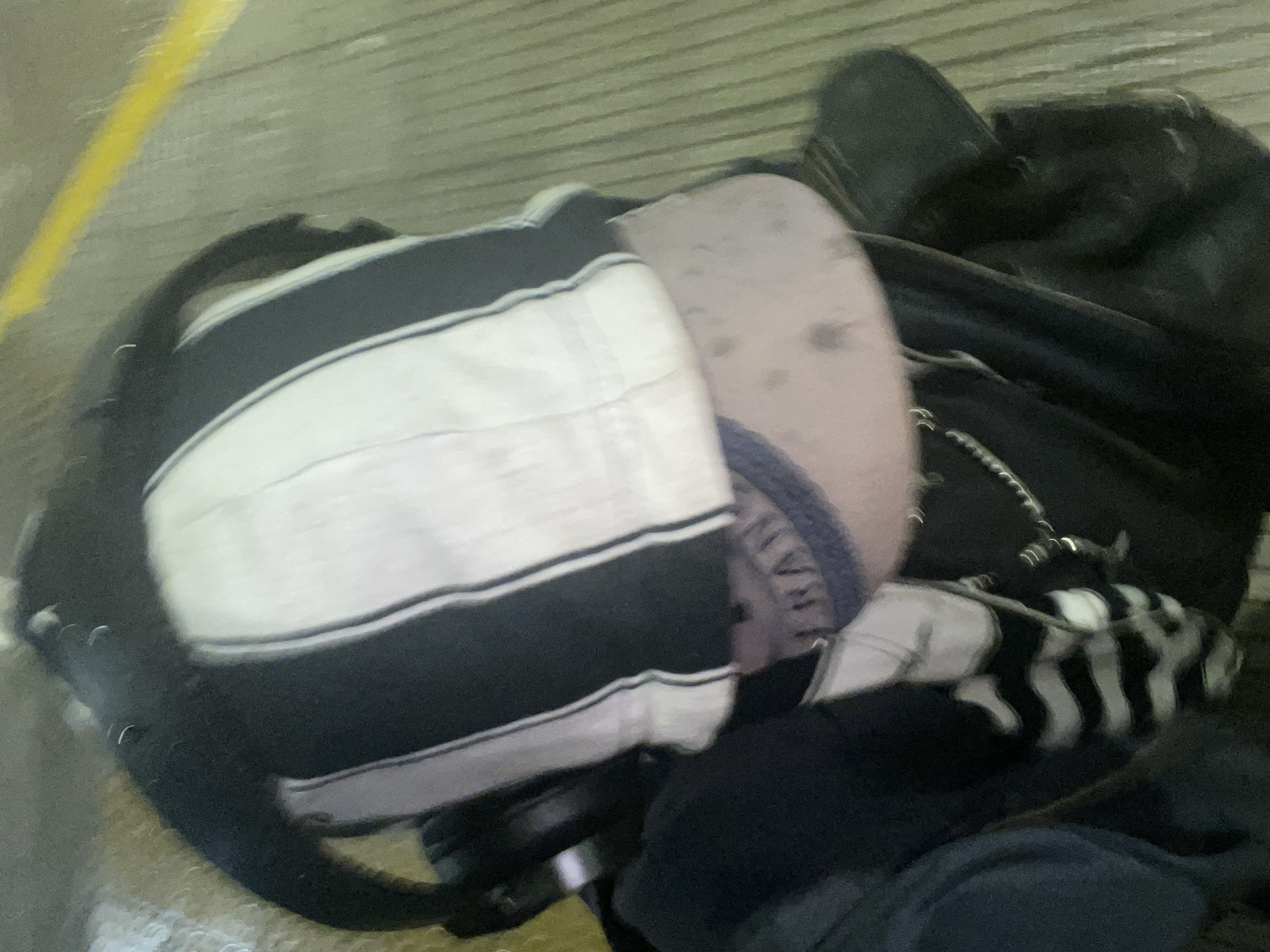 A downward-angled, high-angle view captures the upper back and head of an individual within an indoor public transit environment. The person wears a black and white vertically striped hooded garment. Beneath the hood, a light grey baseball cap, soiled with dark spots and featuring partially legible blue embroidered text (possibly "RAN") on its brim, is worn backward. Dark over-ear headphones are positioned around the neck or atop the striped garment. A silver-colored chain hangs centrally below the cap against a dark garment, potentially a backpack or jacket. The floor is a ribbed, light-colored surface, traversed by a distinct yellow painted line in the upper right quadrant. This setting is consistent with an airport terminal, likely in Buenos Aires, Argentina.