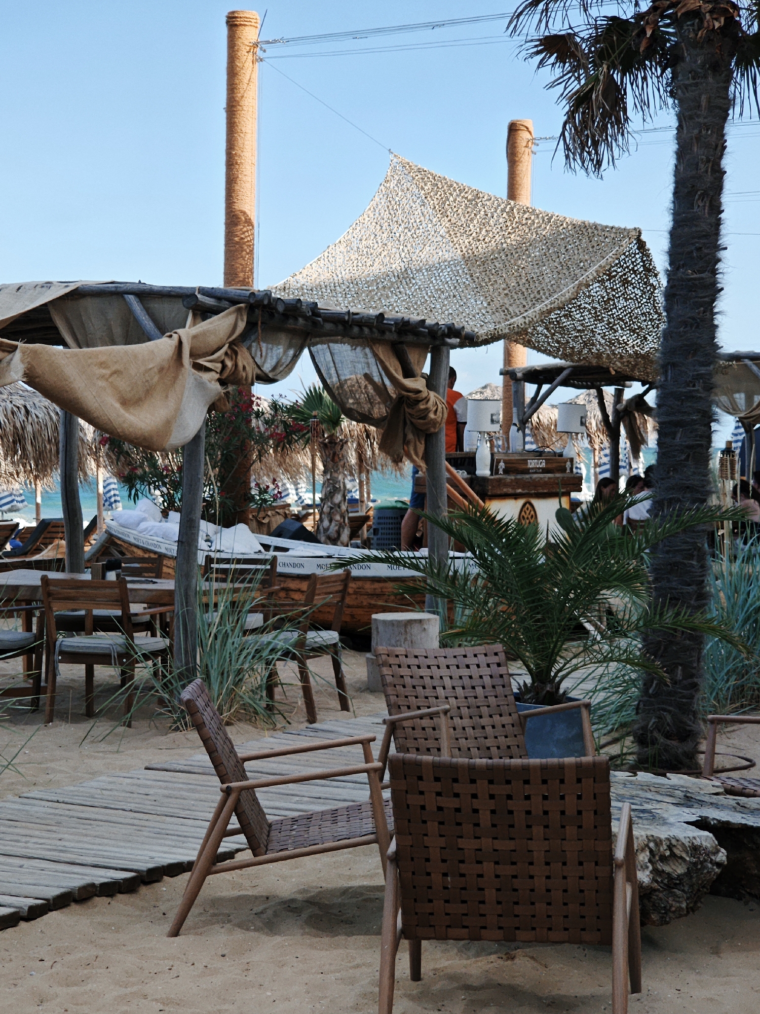 A beachside scene features a covered seating area with woven chairs on a sandy surface. In the background, a boat hull is visible, adorned with the text "MOET & CHANDON." Palm trees and scattered vegetation add to the tropical ambiance, with lounge chairs and umbrellas indicating a resort or dining establishment. The sky is clear and blue, suggesting a sunny day.