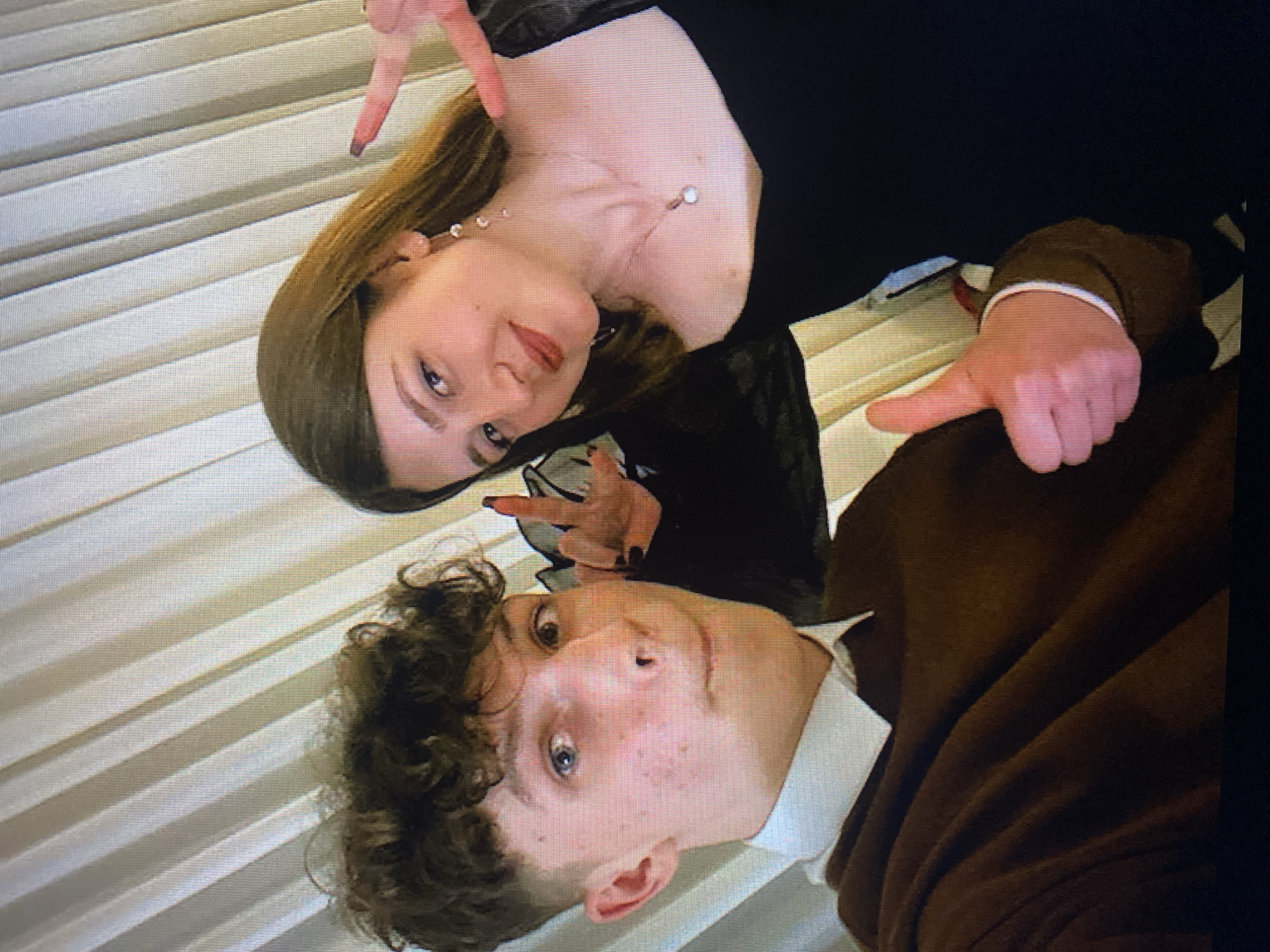 The image shows a close-up selfie of two young people. The person on the left is a young male with curly brown hair, blue eyes, and fair skin with some blemishes. He is wearing a brown sweater over a white collared shirt. His left hand is giving a thumbs-up gesture. The person on the right is a young female with long brown hair, fair skin, and is wearing a black off-the-shoulder dress and a pearl necklace. She is making a peace sign with her right hand and her left hand is partially visible. They are standing in front of a light-colored, vertically striped curtain. The scene appears to be indoors. There is no specific visual information in the image that would allow for identification of the location in the city of Poznan, Poland.