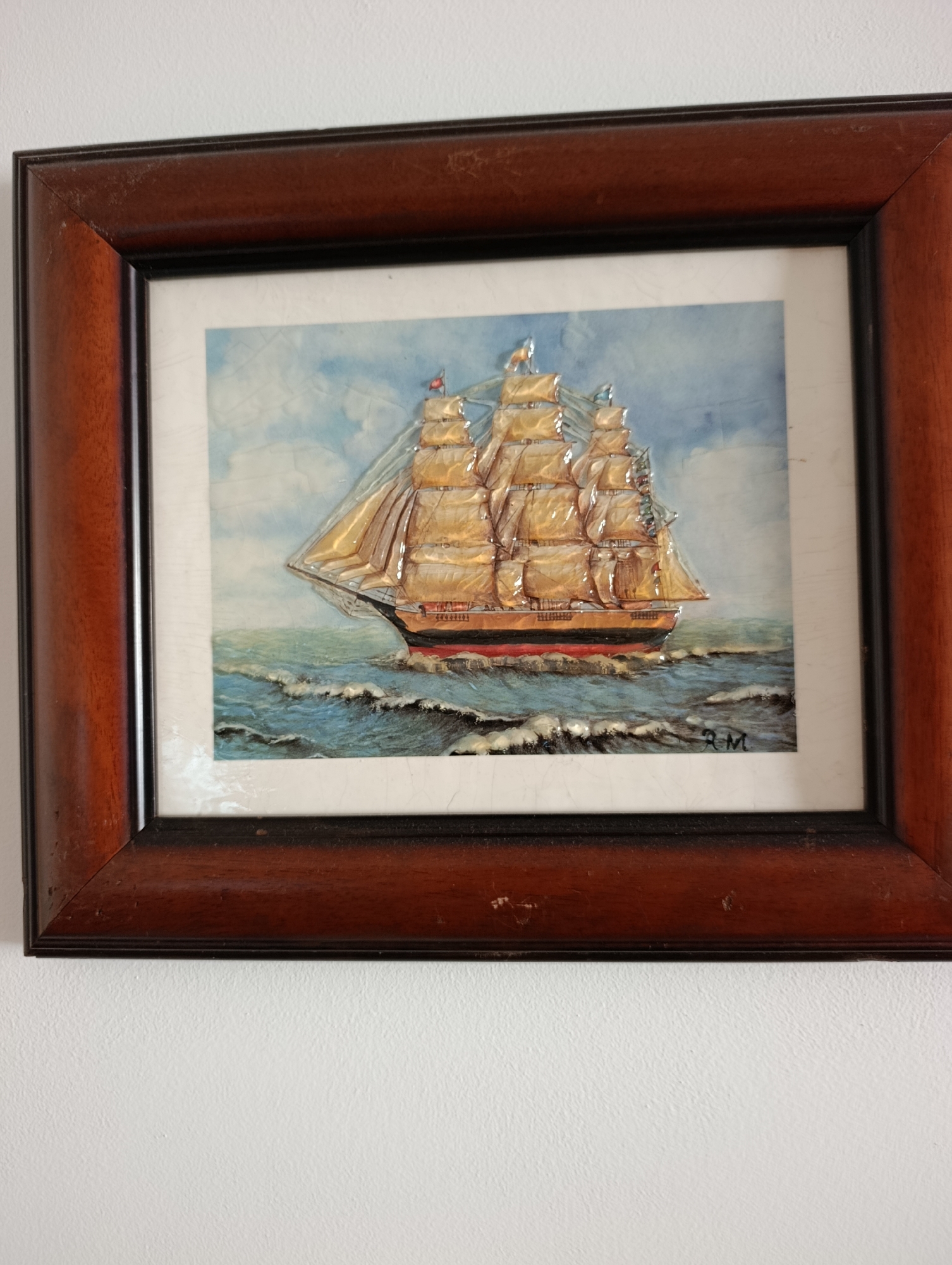 The image shows a framed artwork of a sailing ship on a blue and white background. The ship has three masts and sails that are predominantly a golden-yellow color, with some darker accents. The ship is depicted on choppy blue water with white waves. In the bottom right corner of the artwork, there appears to be a signature or initials. The artwork is housed in a dark brown wooden frame, which shows some signs of wear. The framed artwork is hanging on a textured white wall. The scene is well-lit, likely by natural light. There is no indication of the location in the city of Setúbal, Portugal.