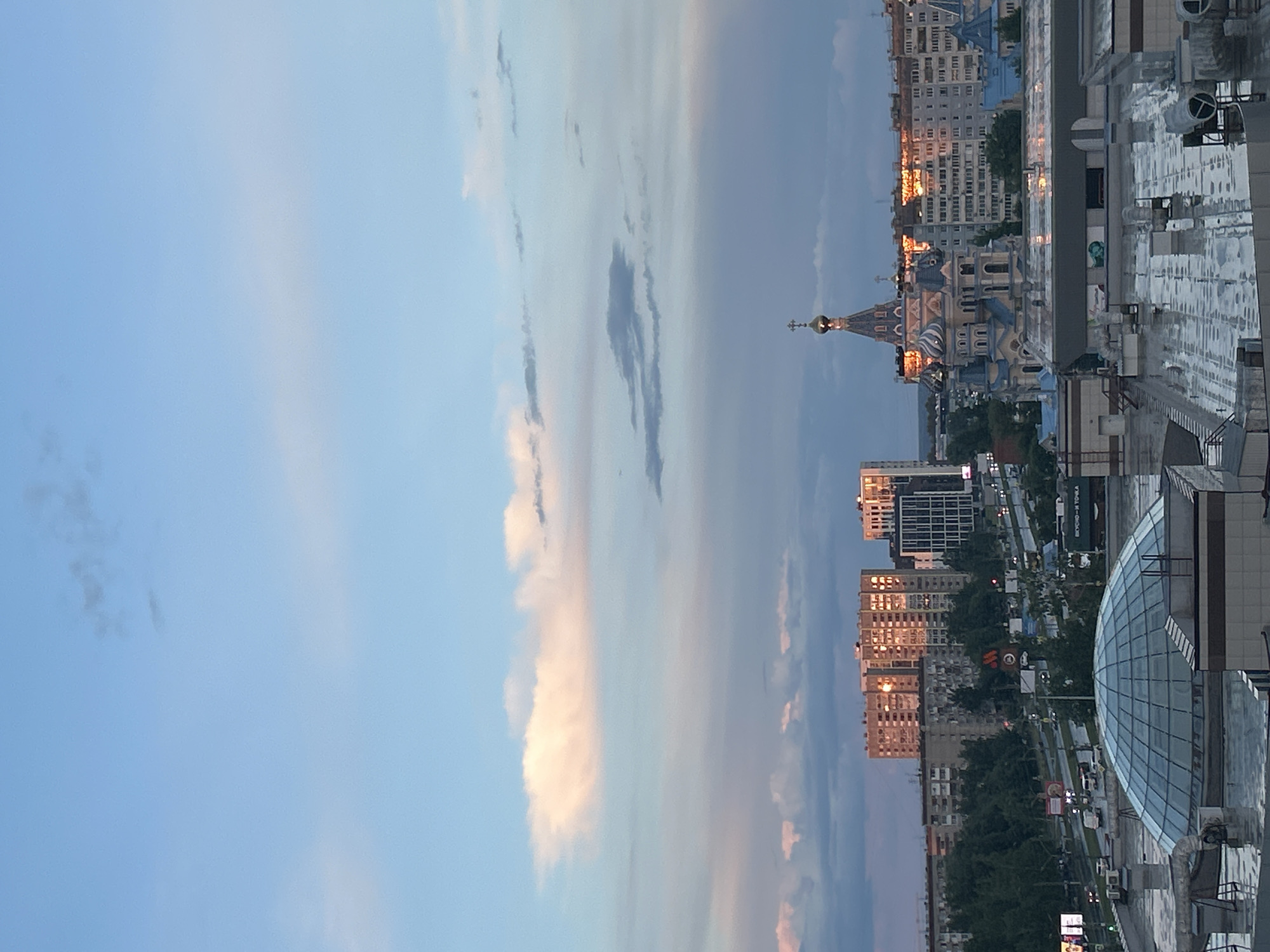 The image shows a skyline view of Izhevsk, Russia, taken from a high vantage point, likely a rooftop. The frame is dominated by a pastel-colored sky with striations of clouds, transitioning from a light blue to darker tones. In the middle ground, various buildings are visible, including residential apartment blocks and taller commercial structures. The most distinctive landmark is the prominent, ornate blue and white Alexander Nevsky Cathedral with its gilded dome. Sun reflects off of a few buildings, suggesting a sunset or sunrise. The foreground features the rooftop from which the image was taken, with visible architectural features like air conditioning units and structures that form the rooftop. The city below appears to be a mix of urban and green spaces, with streets visible between the buildings. The image is oriented vertically, rotated 90 degrees counter-clockwise.