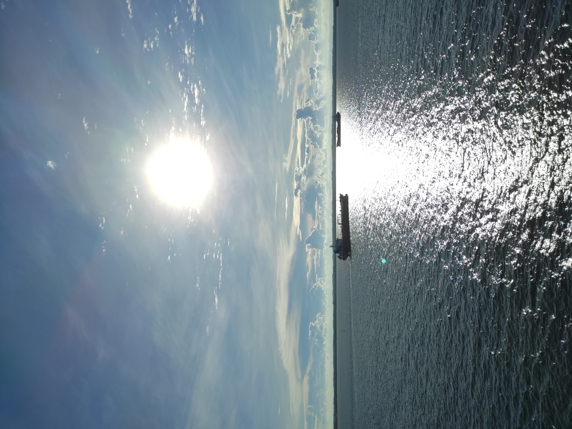 A wide-angle vertical shot captures a bright, sunlit sky over a body of water. The sun, a brilliant white disc with a visible lens flare, dominates the upper center of the frame, surrounded by scattered white clouds and streaks of cirrus clouds against a light blue sky. A shimmering reflection of the sun stretches across the dark, choppy water in the foreground.
On the horizon, a low, distant shoreline is visible. Two cargo ships are present in the water. One, a dark-hulled vessel, is positioned in the middle ground towards the left side of the frame, appearing to be a tanker. Further to the right, a second, smaller dark vessel is visible. The water is a deep blue-grey, with whitecaps and ripples indicating movement. The overall impression is one of a sunny day on a wide expanse of water, likely a bay or strait, with maritime activity. The scene is consistent with a view from the coast or a vessel in the waters near Penajam, Indonesia, given the typical maritime traffic and coastal geography of the area.