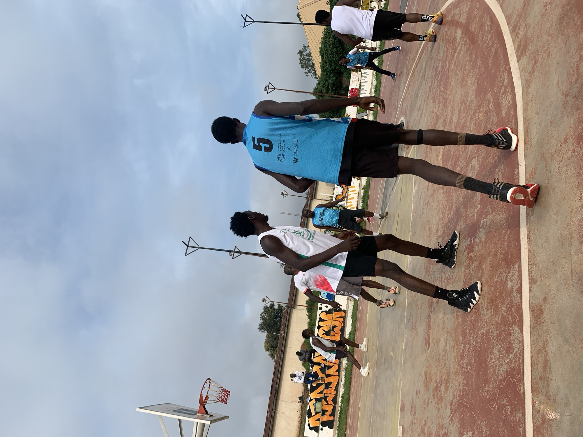 The image depicts an outdoor basketball court in Gura Top, Nigeria, under a cloudy sky. Several men are engaged in a game of basketball. In the foreground, a man in a blue jersey with the number "5" is facing away from the camera, wearing black shorts and black socks with brown bands. To his left, another man in a white jersey with green trim and black shorts is in a defensive stance. In the background, other players are scattered across the court. A basketball hoop with a red net is visible on the left side of the image, and a wall with colorful graffiti runs along the back of the court. The court surface is red with white lines.